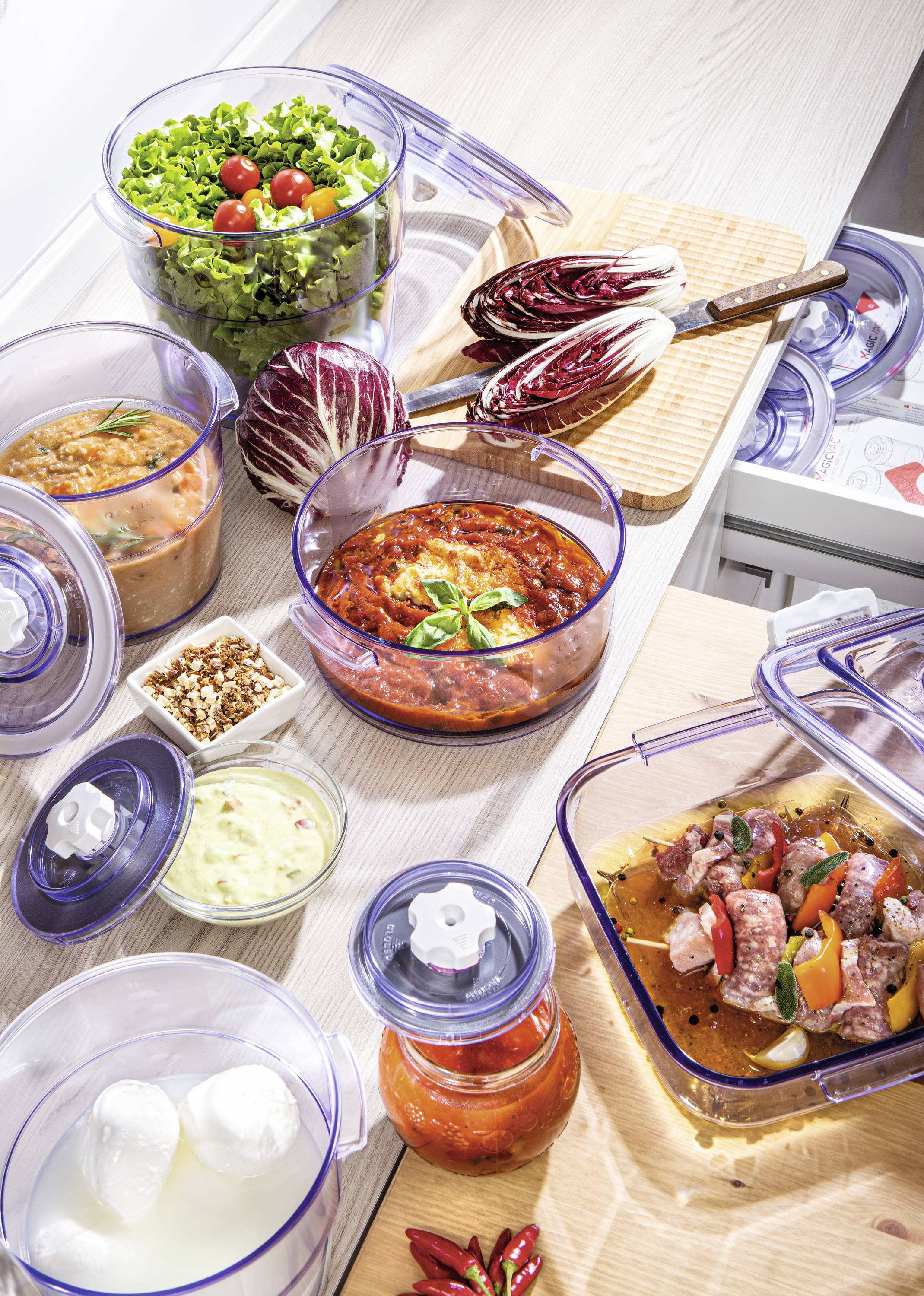 Čerstvé ingredience a připravené pokrmy v průhledných nádobách, včetně salátu, rajčatové omáčky a masa, na kuchyňské pracovní desce.