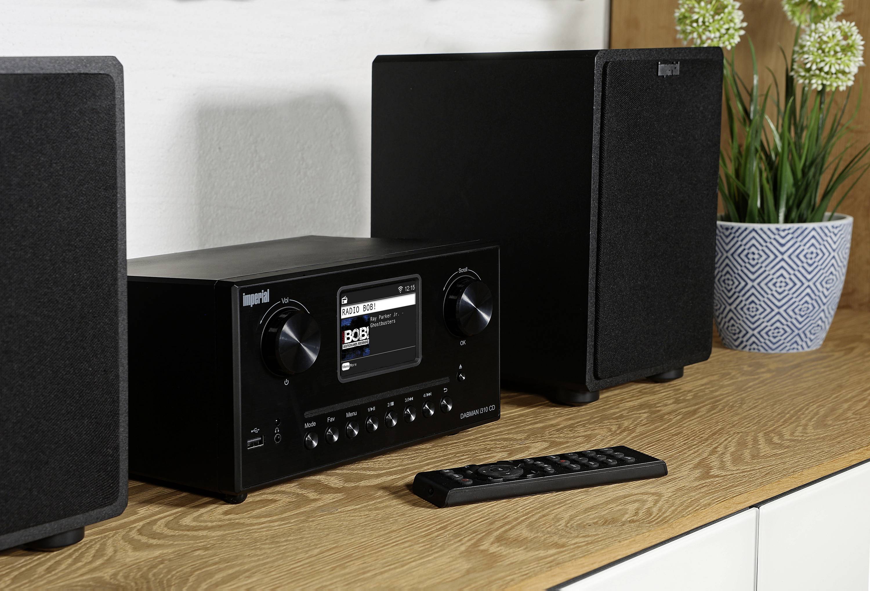 Černá stereosoustava s digitálním displejem a dvěma reproduktory na dřevěném stole, vedle ní dálkový ovladač a rostliny v pozadí.