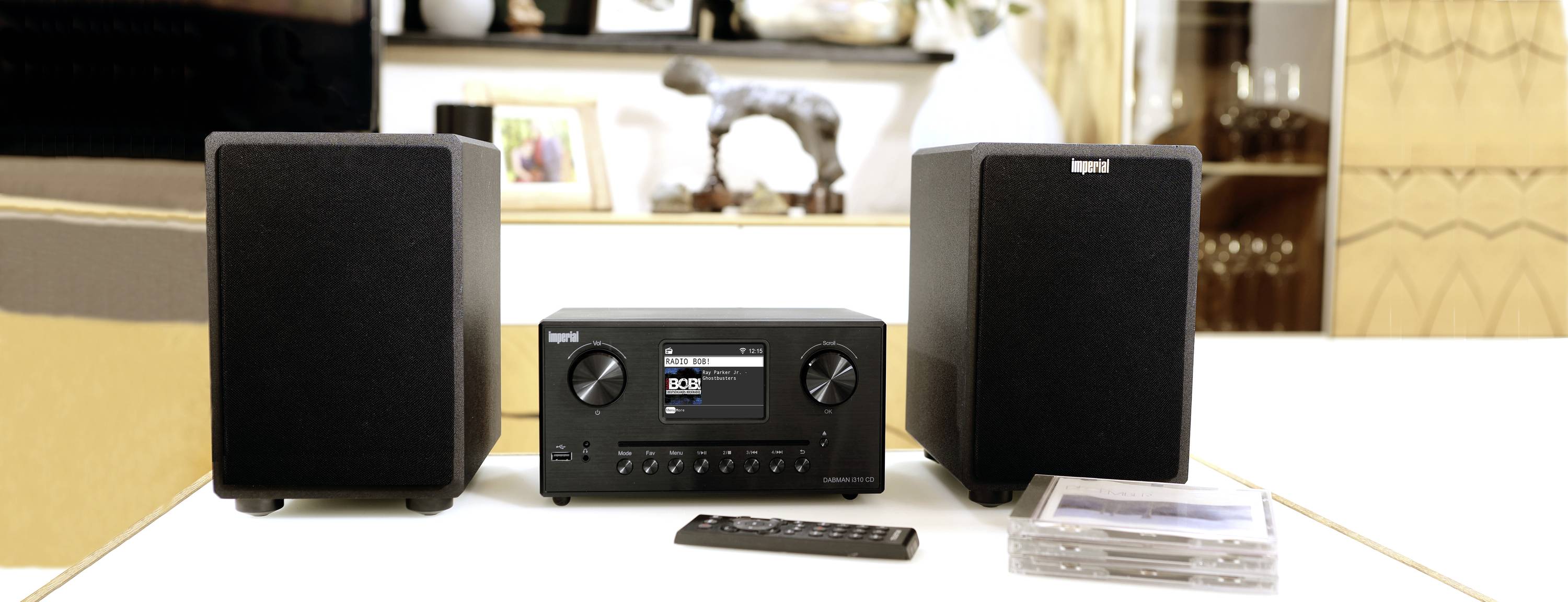 Černý stereofonní audiosystém se dvěma reproduktory, hlavní jednotkou s displejem a otočnými ovladači; dálkový ovladač a CD disky vedle něj.