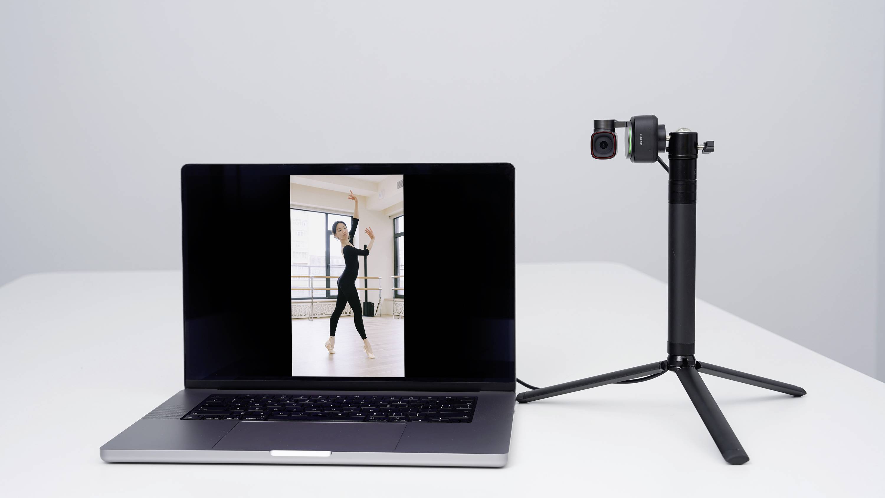 Laptop zobrazuje obrázek tančící ženy. Vedle něj stojí fotoaparát na stativu, který je namířen na laptop.