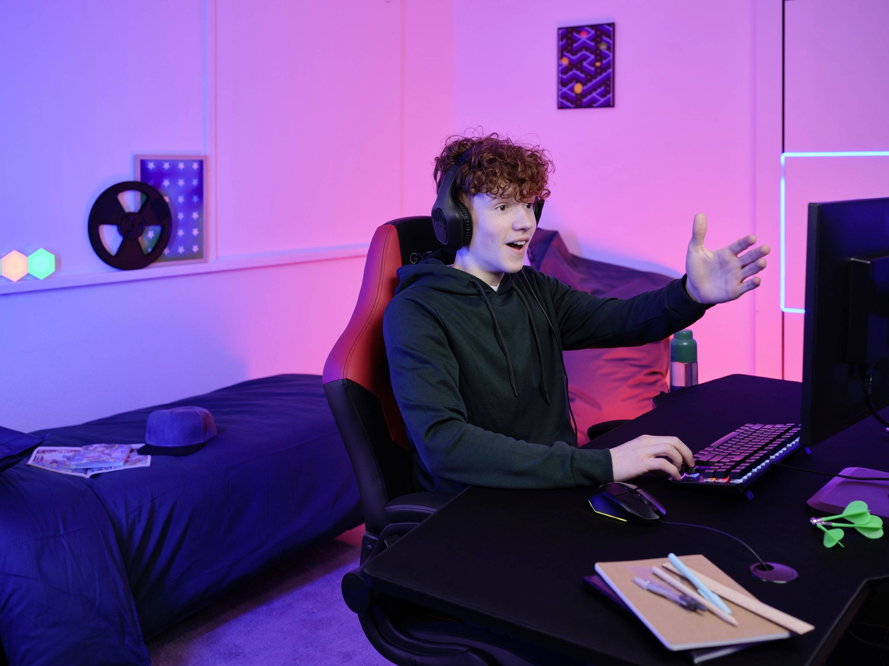 Mladý muž sedí u psacího stolu a hraje na počítači, má nasazené sluchátka. V pozadí je vidět postel.