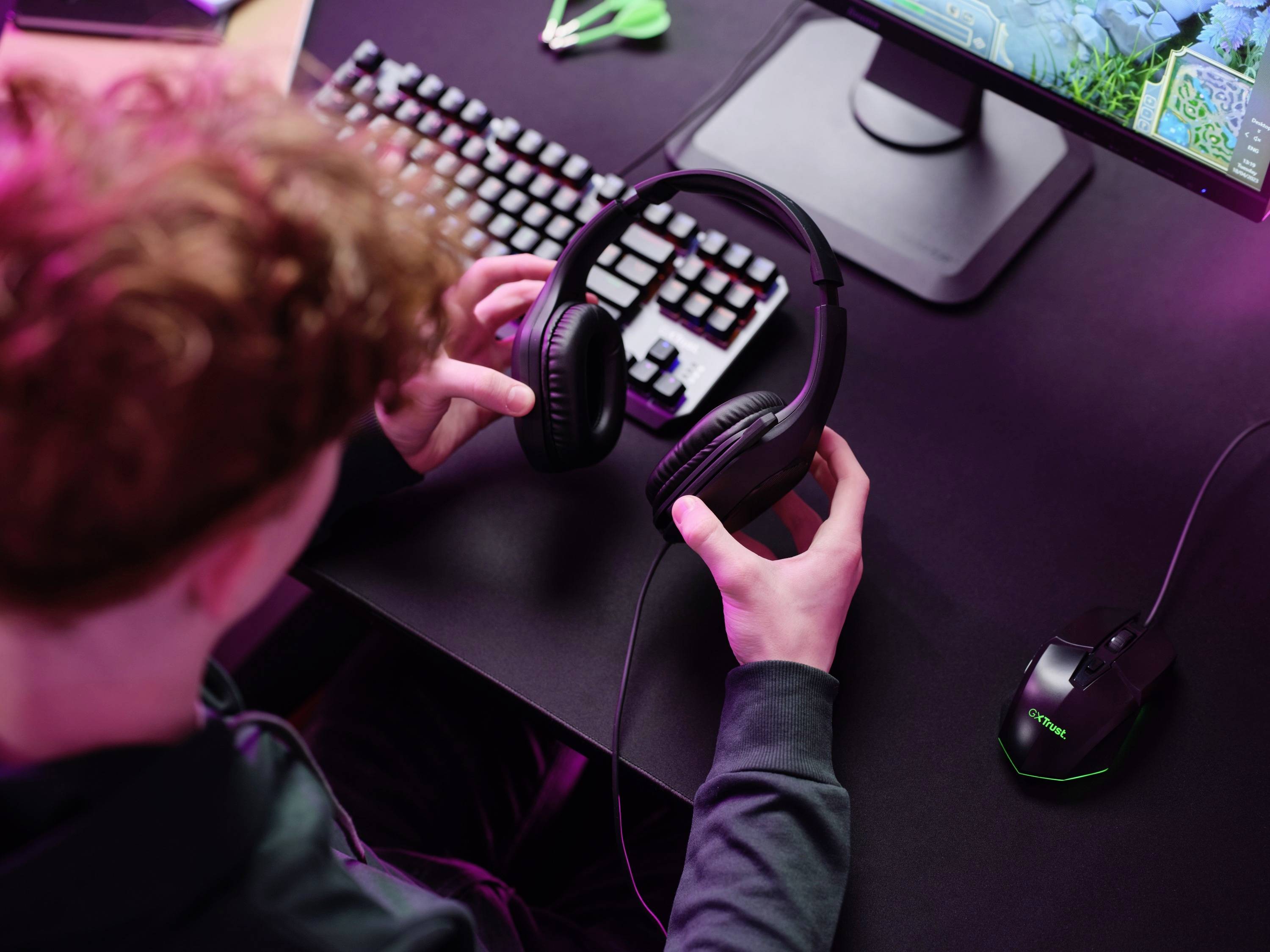Osoba sedí u stolu s počítačem a drží v rukou headset. Je vidět barevná klávesnice a myš.