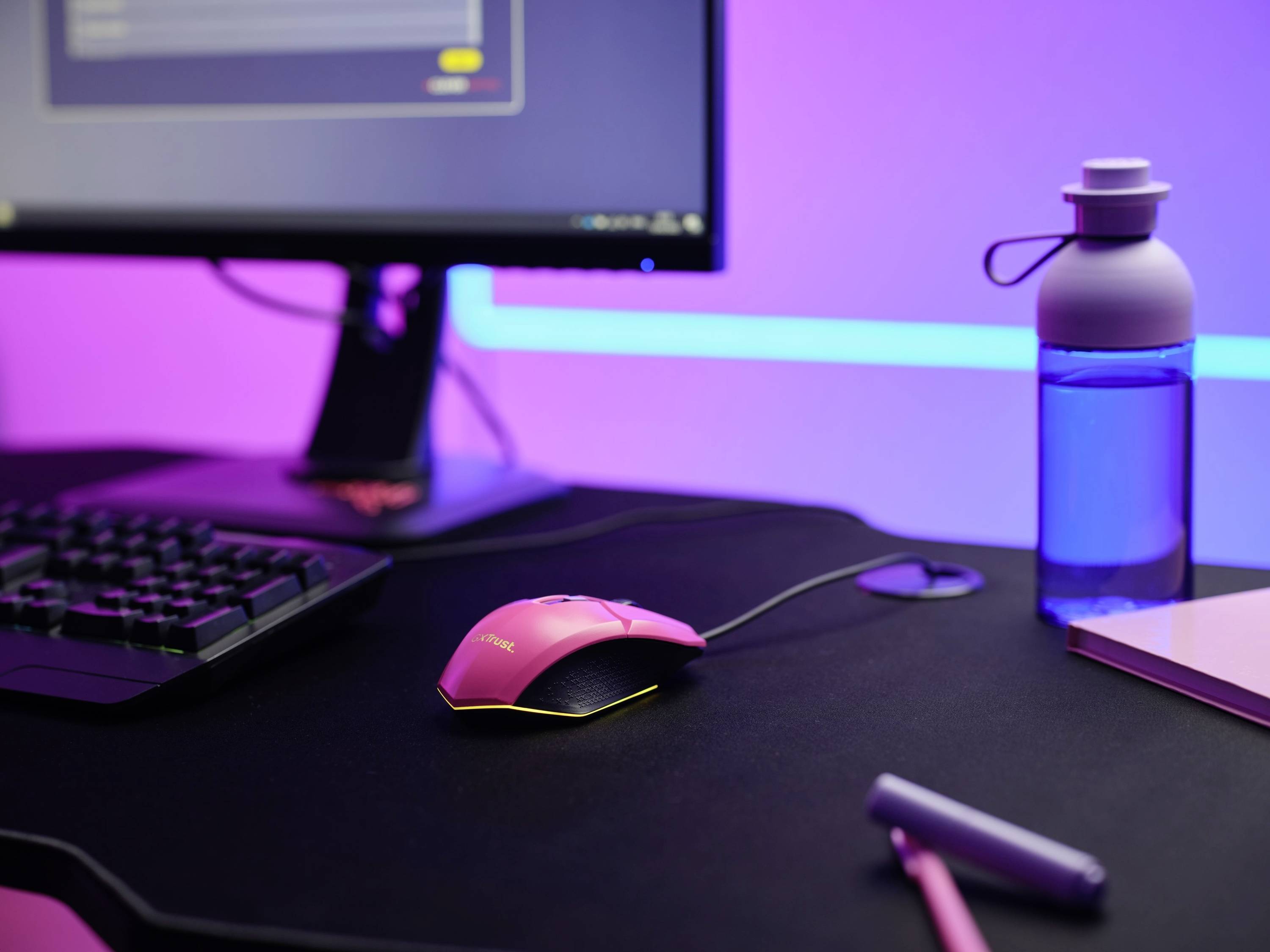 Psací stůl s růžovou počítačovou myší, klávesnicí, monitorem, modrou láhví na vodu a růžovým blokem.