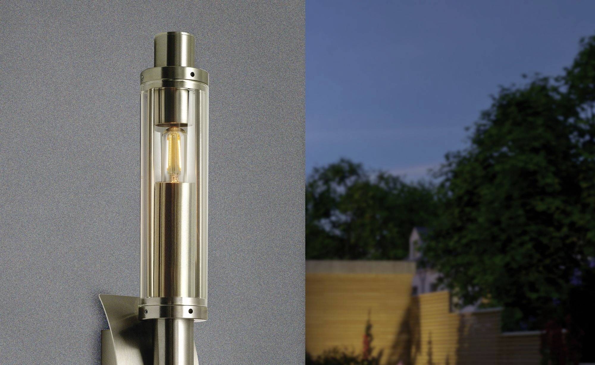 Moderní venkovní lampa osvětluje zeď. V pozadí jsou rozmazané stromy a dřevěný plot za soumraku.