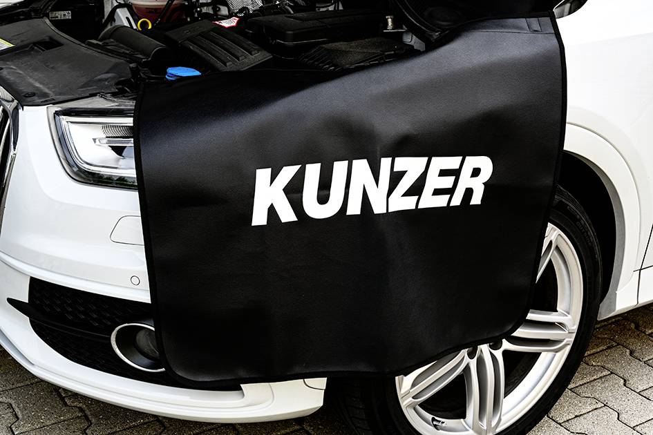 Bílé auto s otevřenou kapotou motoru, zakryté černou ochrannou podložkou s nápisem 'KUNZER'.