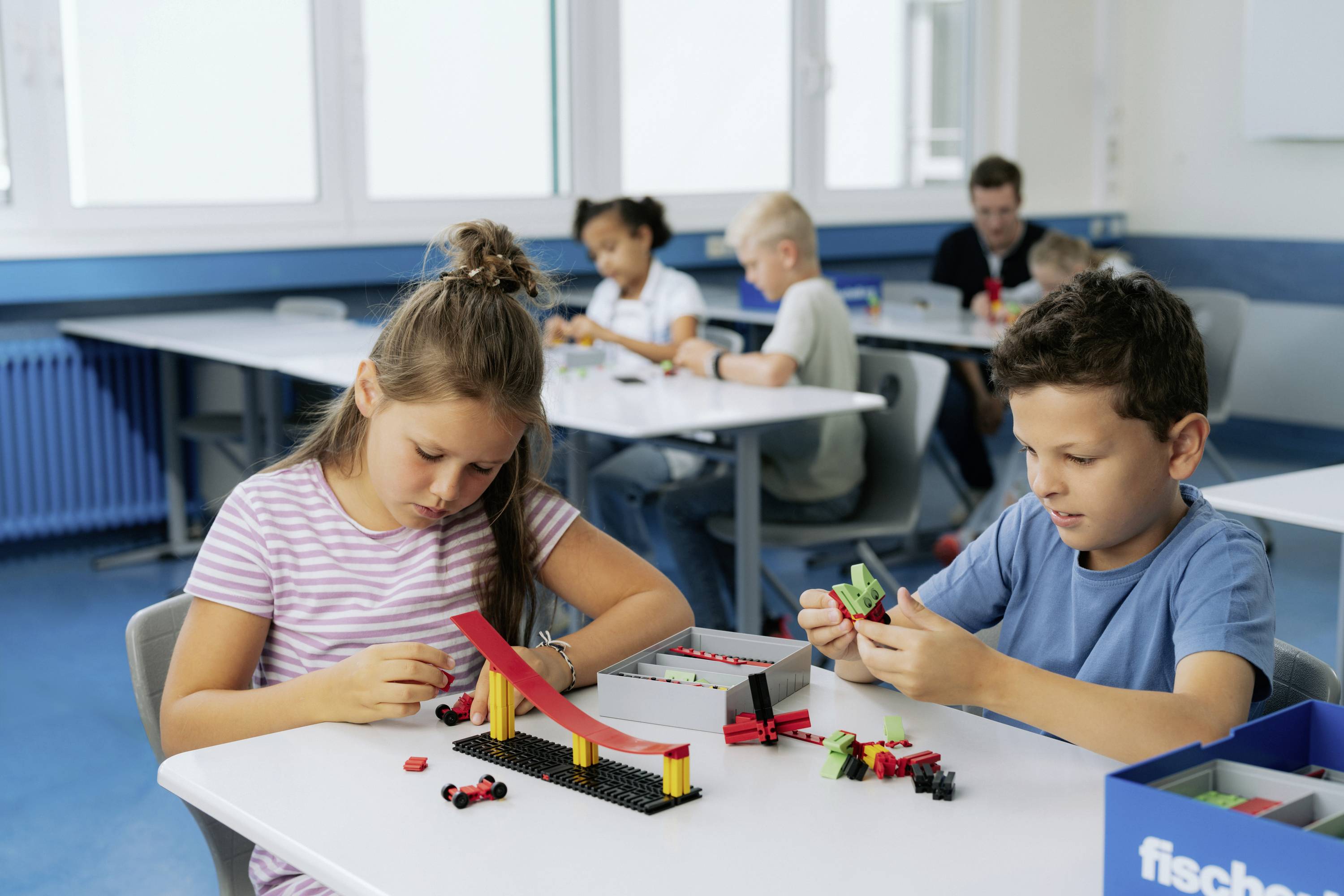 Děti staví z kostek v učebně. Několik stolů, u kterých žáci pracují. Důraz je kladen na kreativitu a spolupráci.