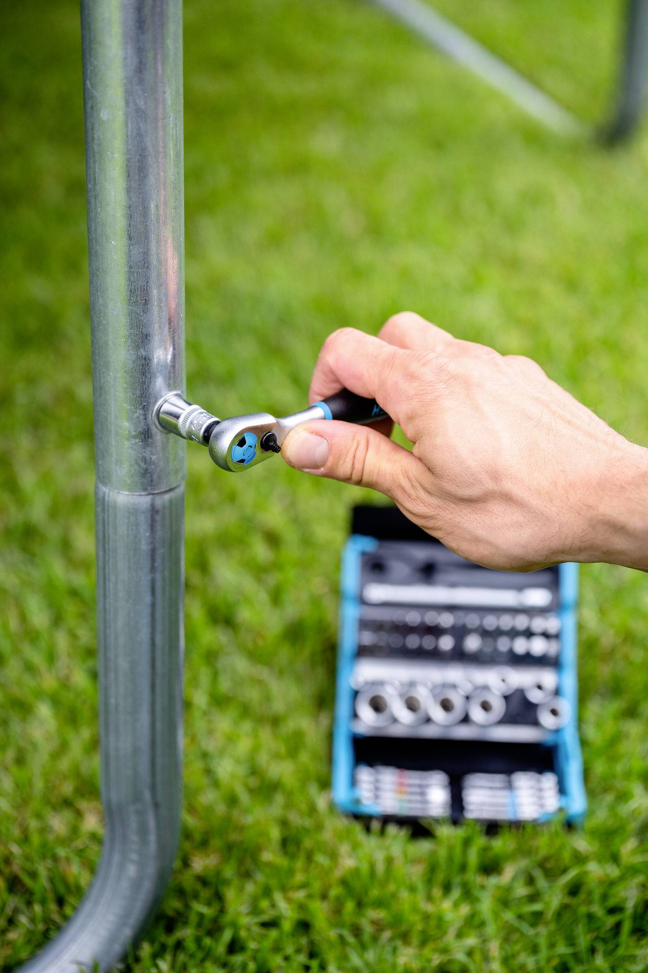 Osoba utahuje šroub pomocí ráčnového klíče na kovovém potrubí venku; níže na trávě je viditelný montážní kufřík s různými nástroji.