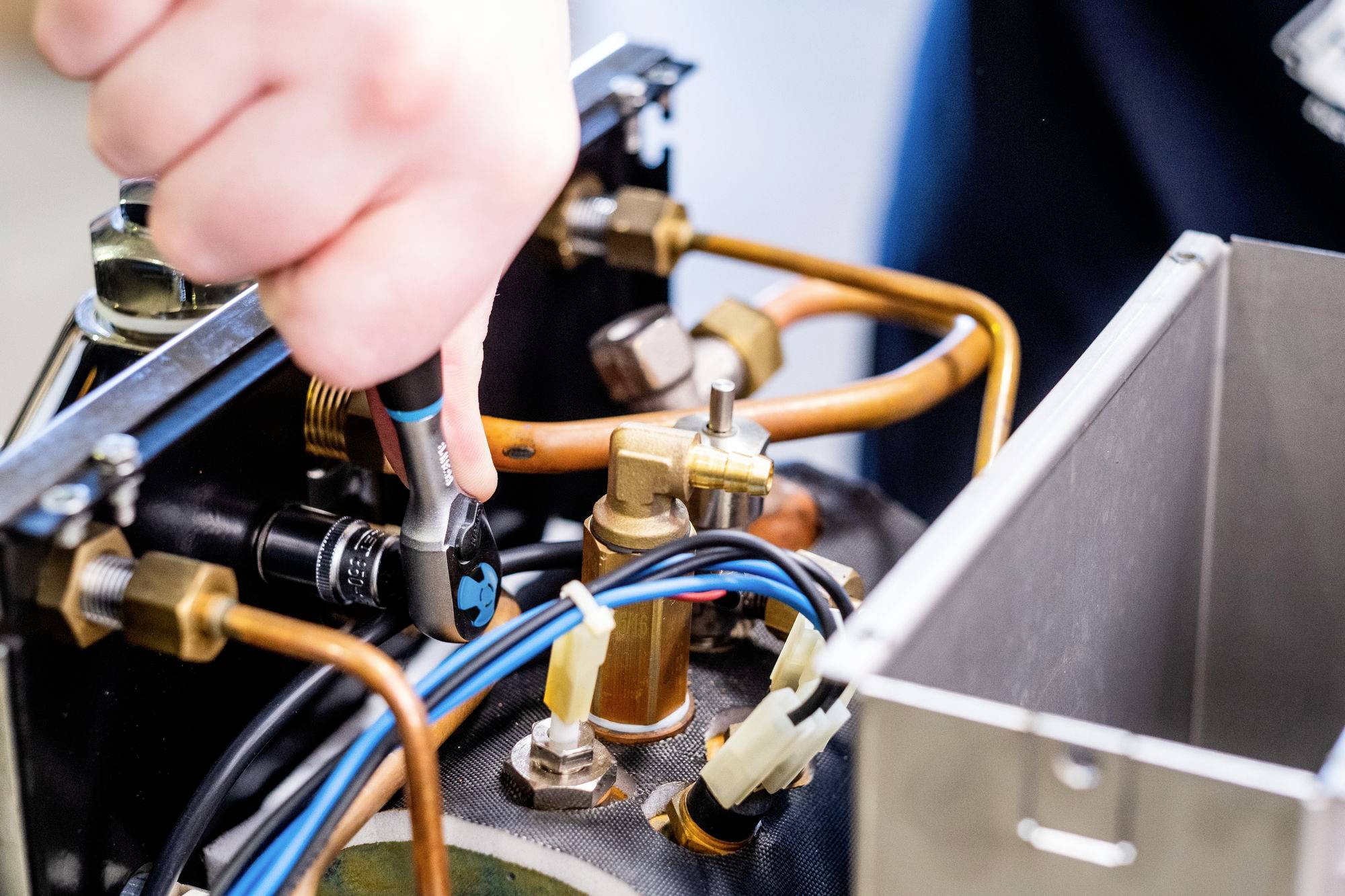 Osoba používá ráčnový klíč k utažení kovového komponentu mezi měděnými trubkami a elektrickými vodiči uvnitř mechanického zařízení.