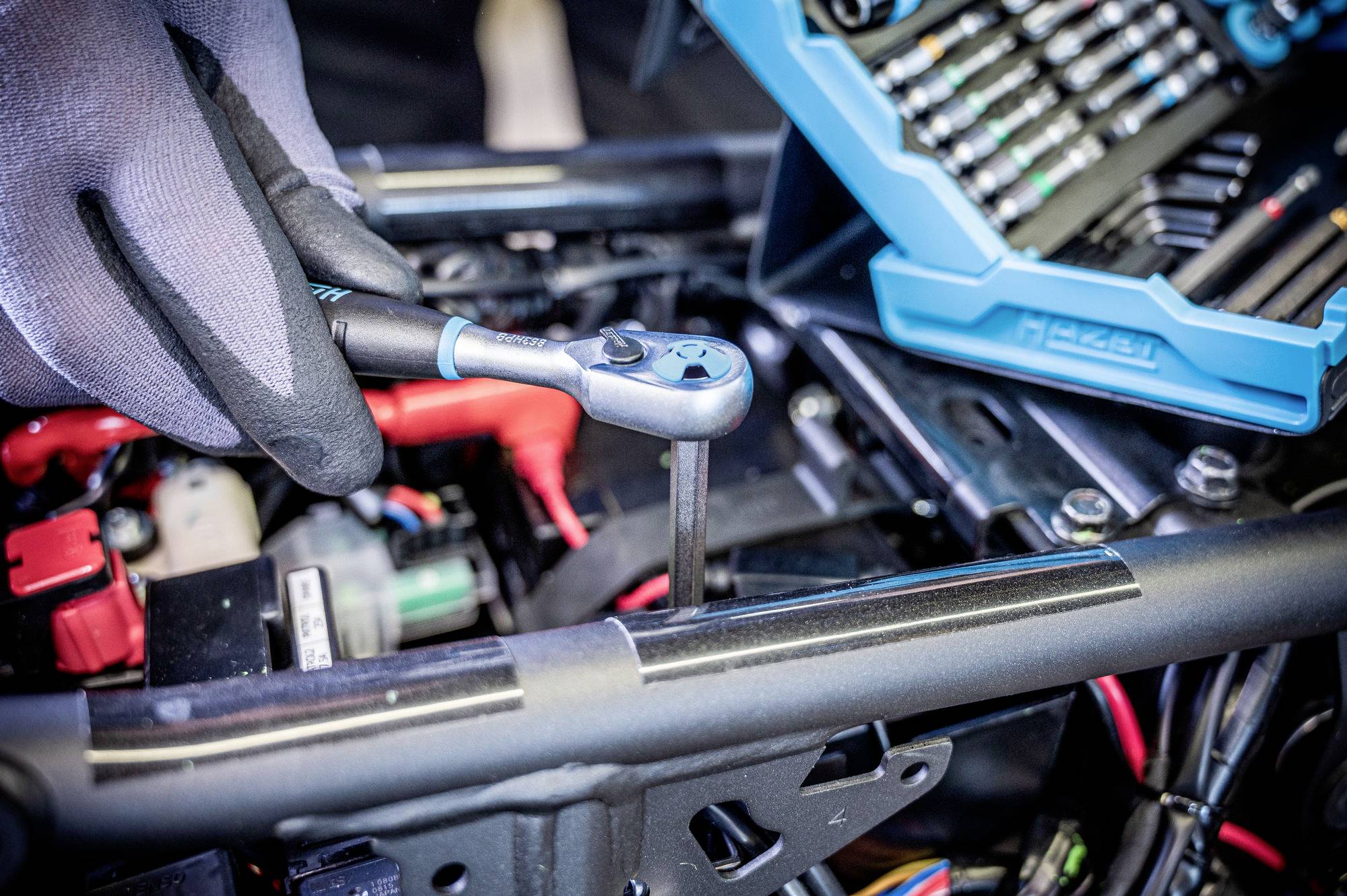 Osoba v rukavicích používá ráčnový klíč k utažení šroubu na mechanickém zařízení, přičemž v pozadí je viditelná montážní bedna.