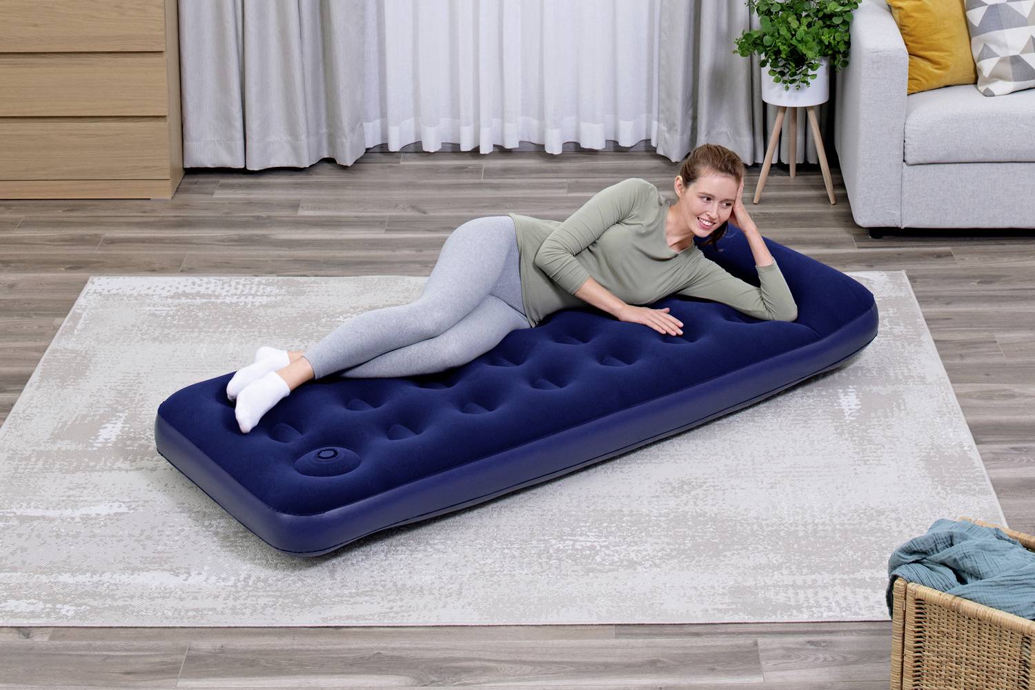 Osoba relaxuje na modré nafukovací matraci v obývacím pokoji. V pozadí jsou vidět záclony a pohovka.