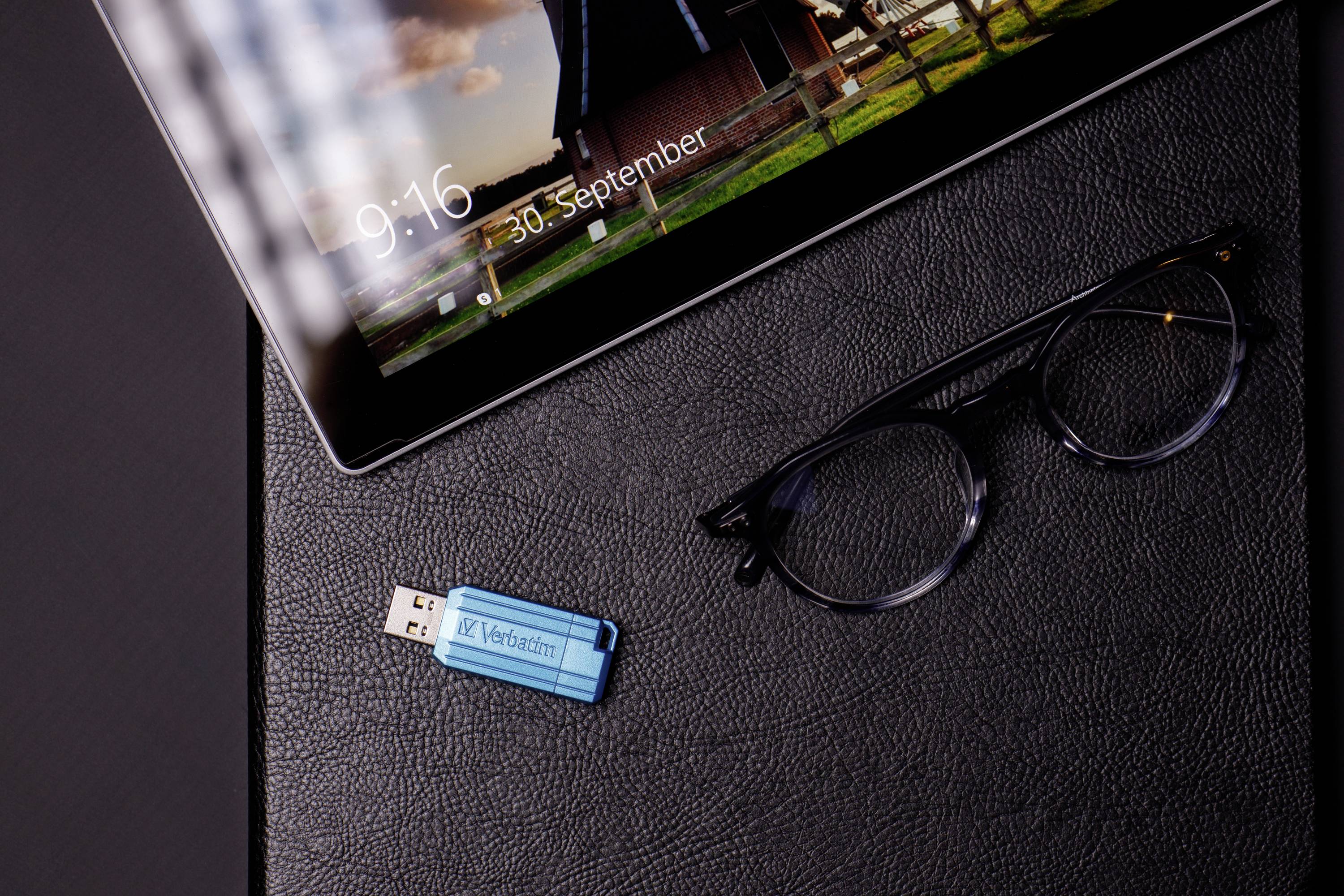 Tablet s uzamčenou obrazovkou zobrazuje '9:16 30. září'. Vedle něj leží brýle a modrý USB flash disk na tmavém povrchu.