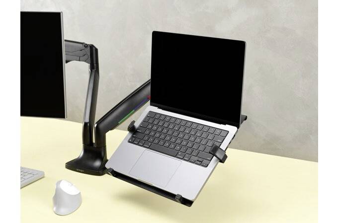 Laptop leží na nastavitelném stojanu vedle monitoru s ramenem a počítačovou myší na světlém psacím stole.