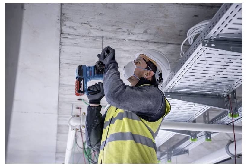 Dělník v ochranné přilbě a reflexní vestě používá elektrickou vrtačku na stropě, obklopen elektrickými vodiči a trubkami.
