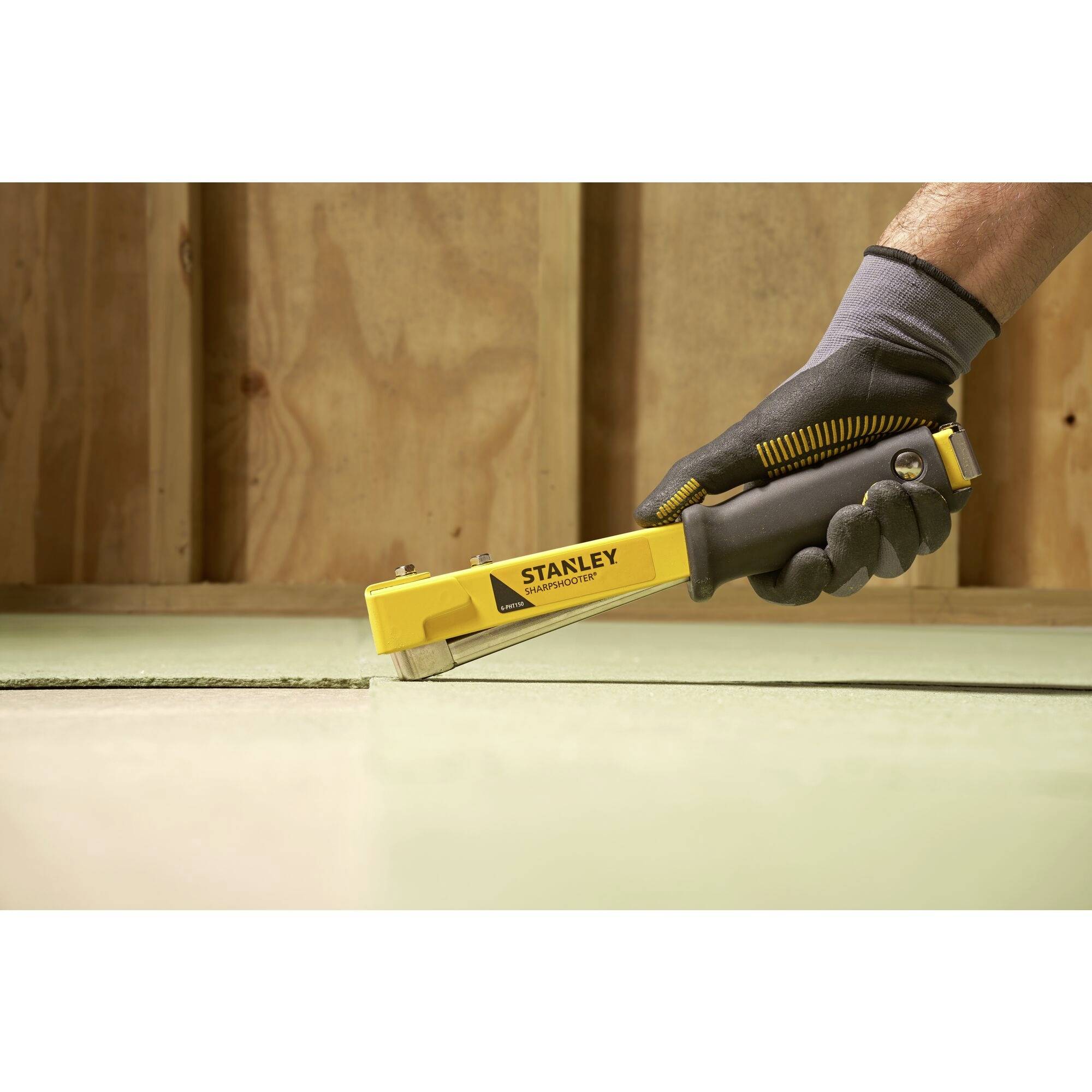 'Close-up of a gloved hand operating a yellow Stanley stapler. The stapler is used to fasten material to wood.'