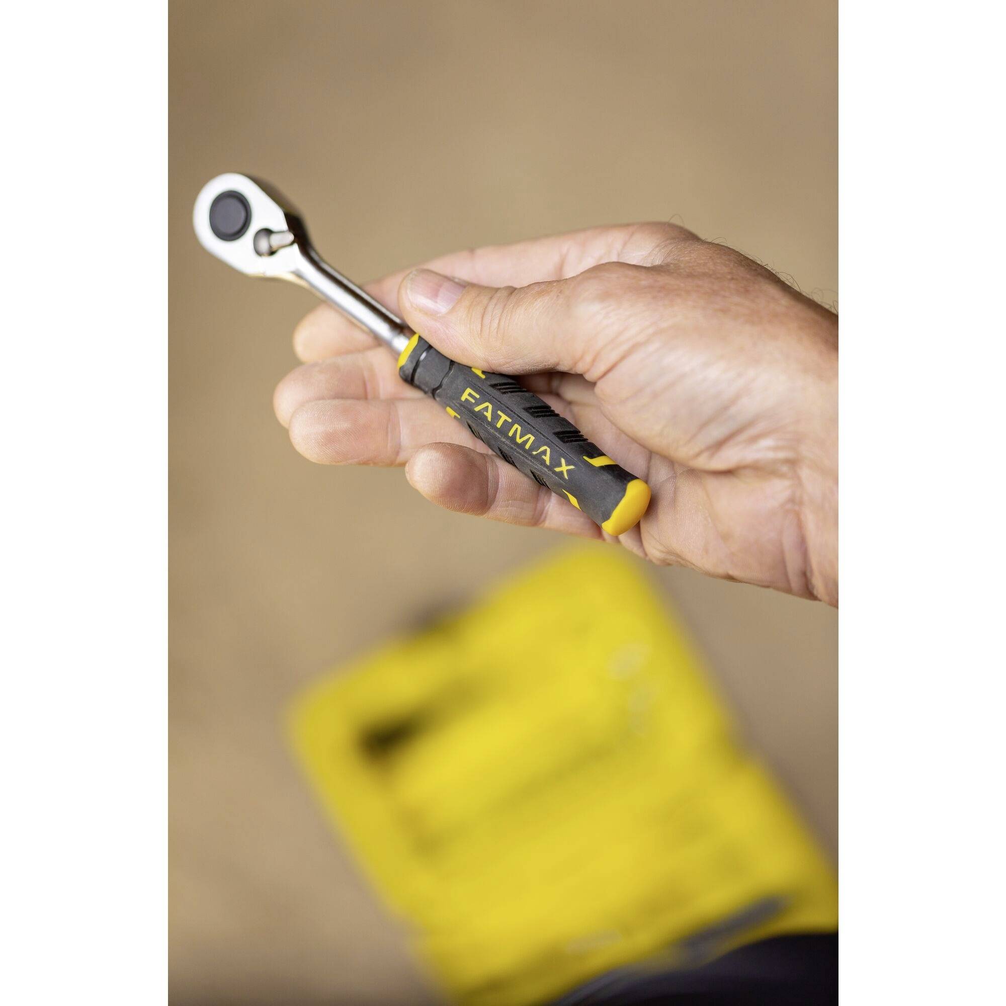 Ruka drží ráčnový klíč s černo-žlutou rukojetí. V rozostřeném pozadí je viditelná brašna na nářadí.