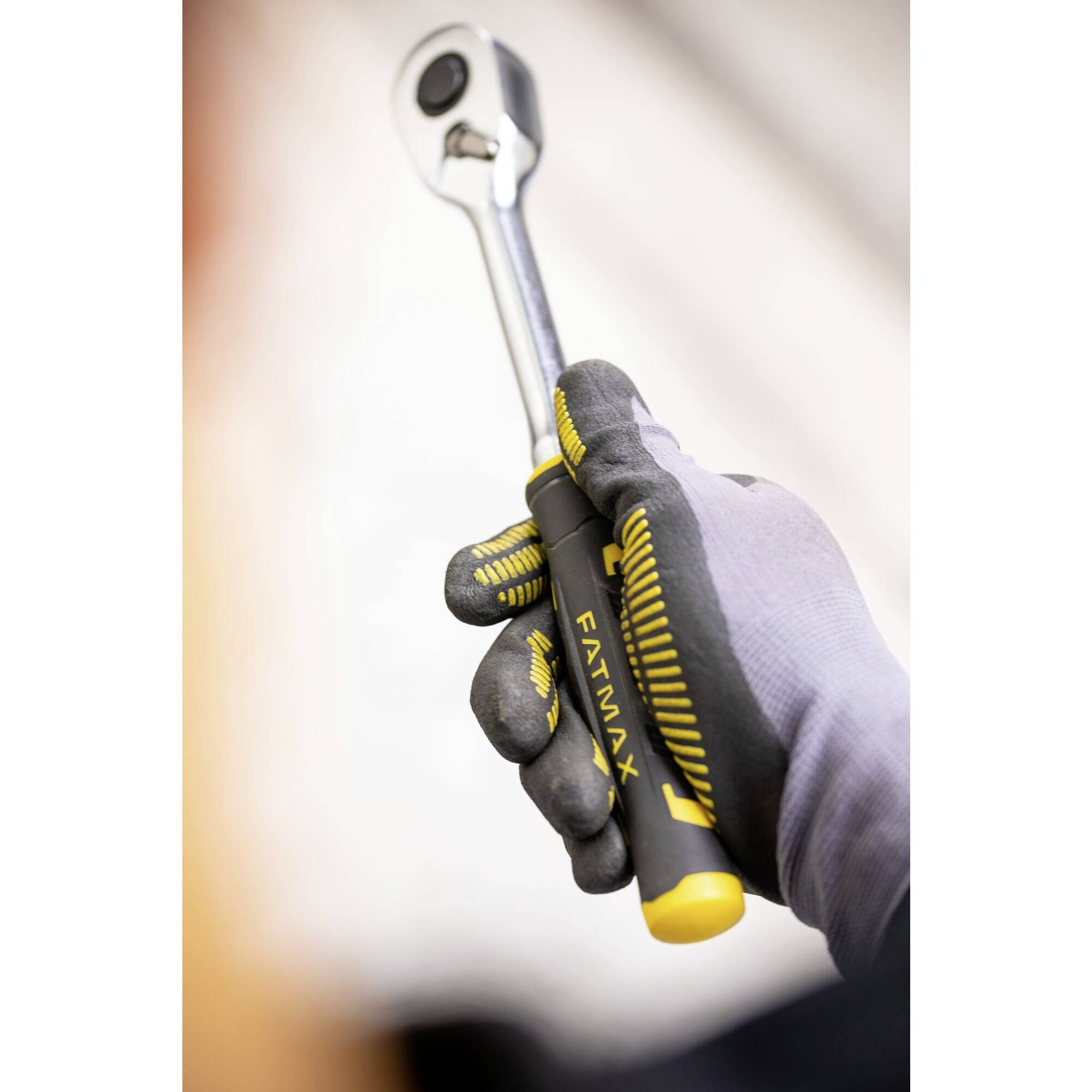 Ruka v šedé rukavici drží ráčnový klíč s černou a žlutou rukojetí.