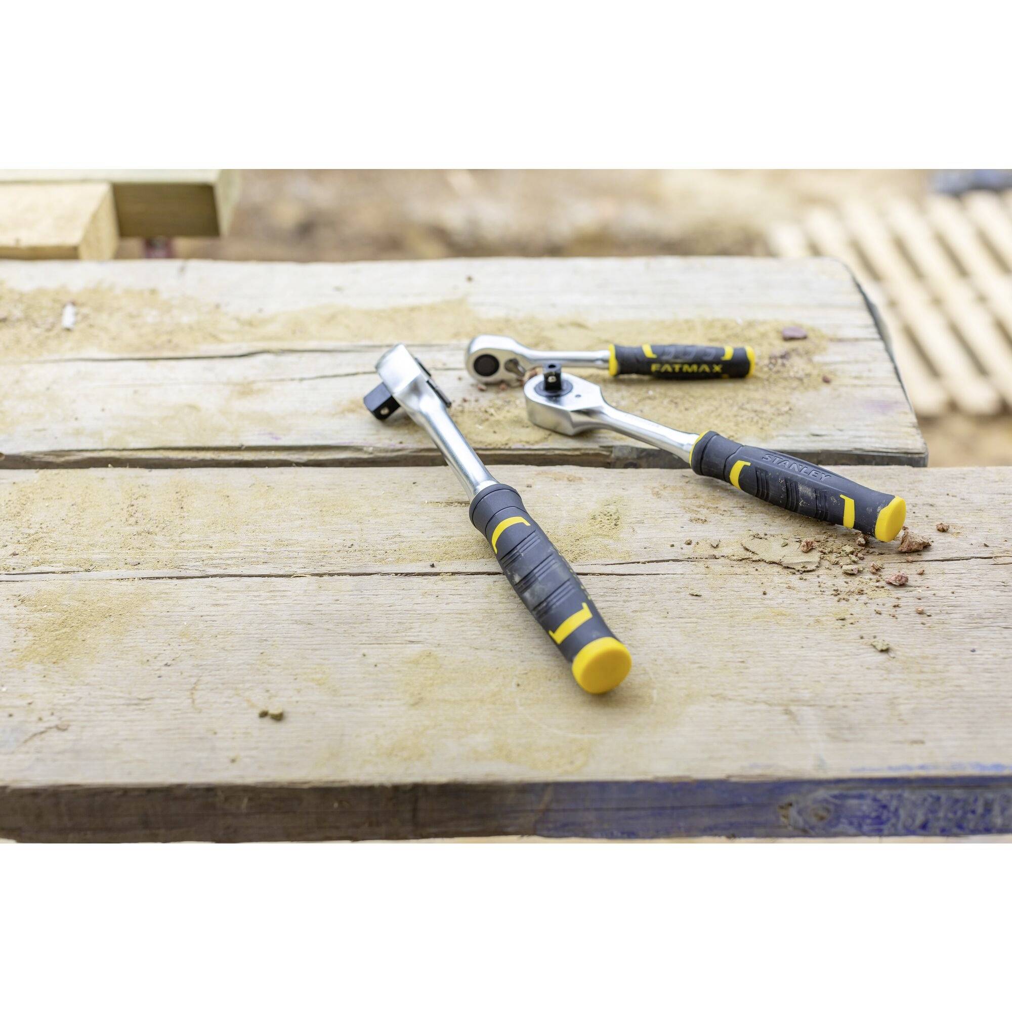 Na dřevěném stole leží dvě ráčny se žlutými rukojetmi; jedna je větší než druhá. V pozadí jsou vidět dřevěné desky.