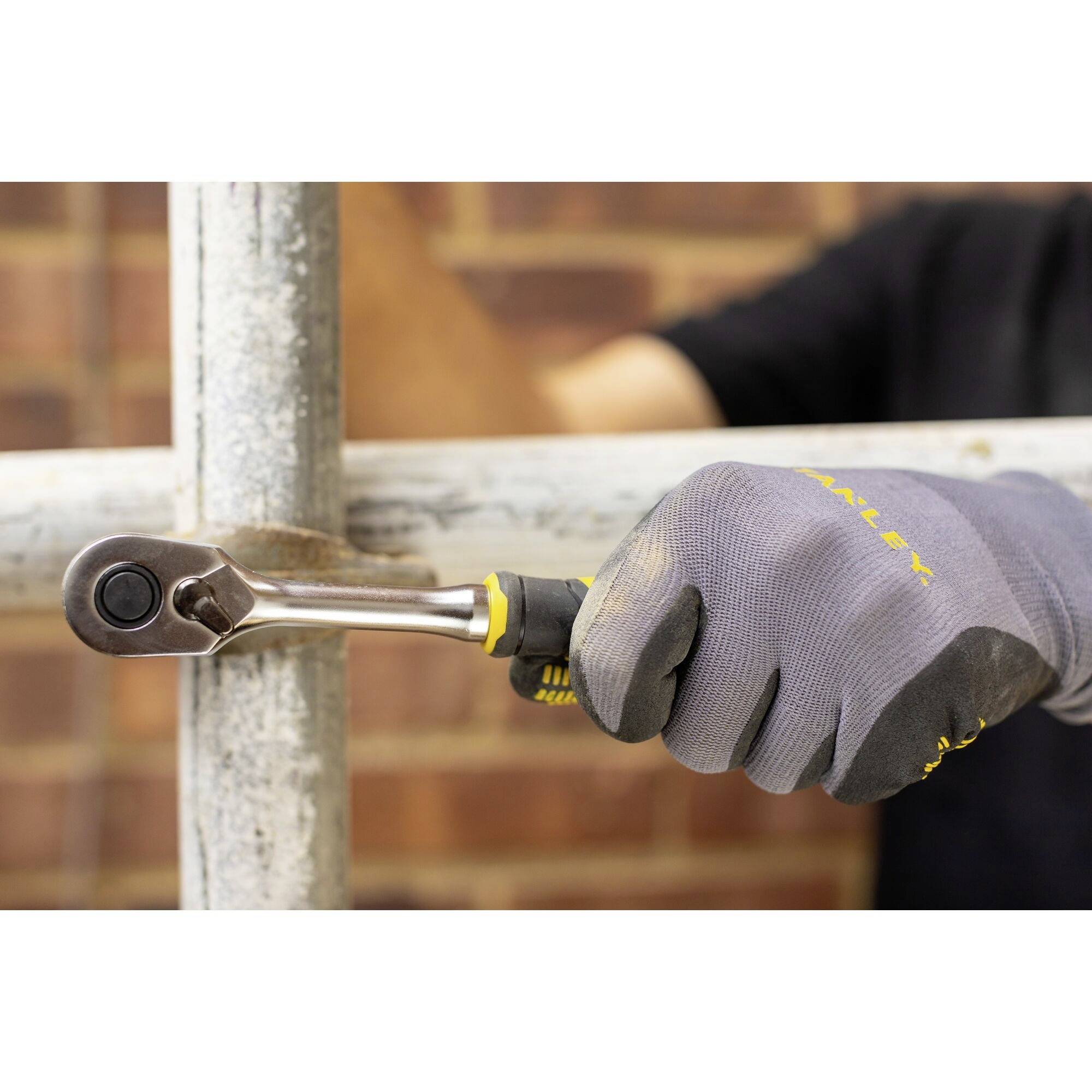 Ruka v rukavici drží nástrčný klíč a utahuje šroub na kovovém potrubí. V pozadí je rozostřená stěna.