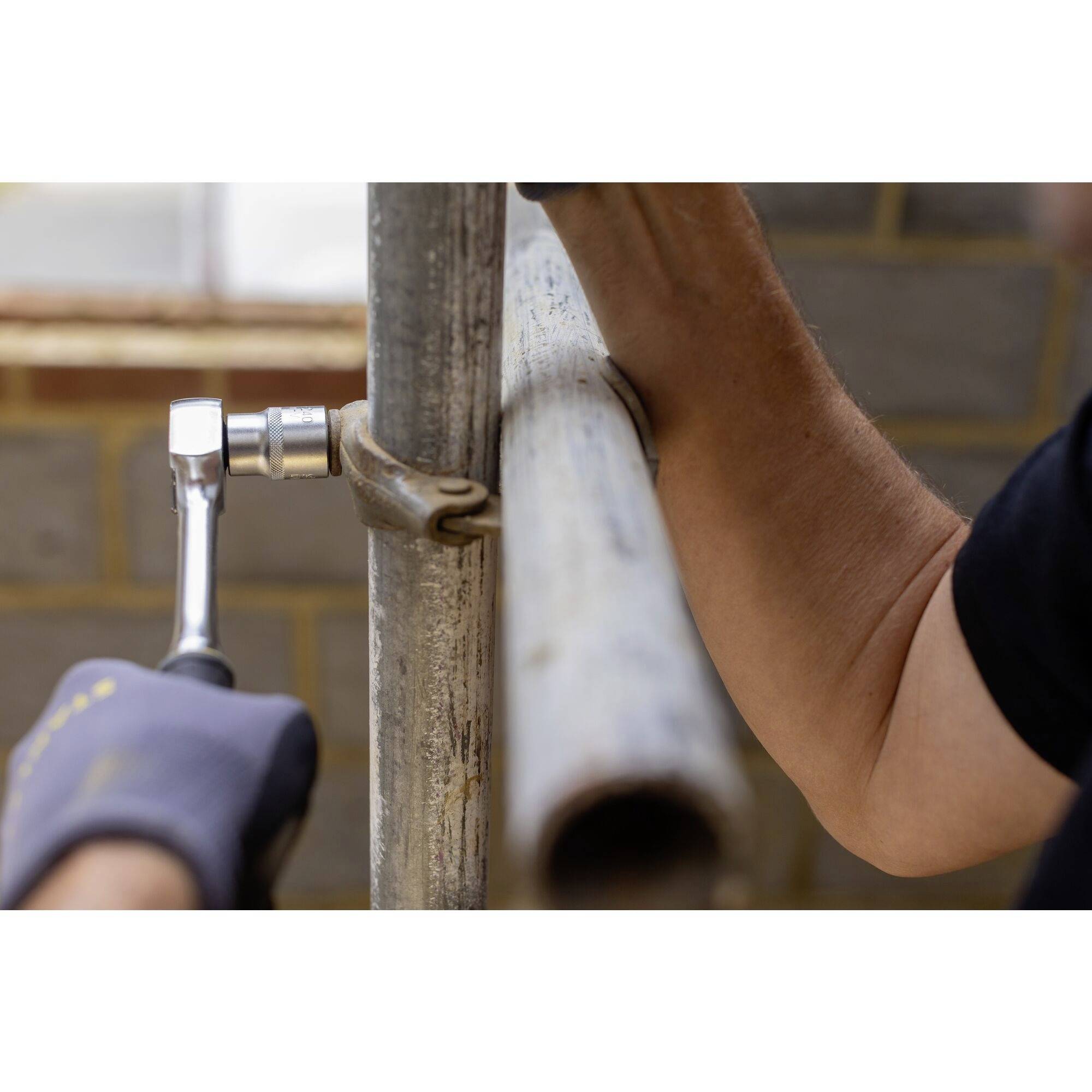 Osoba utahuje svorku na kovovém potrubí pomocí klíče. V pozadí je cihlová zeď.