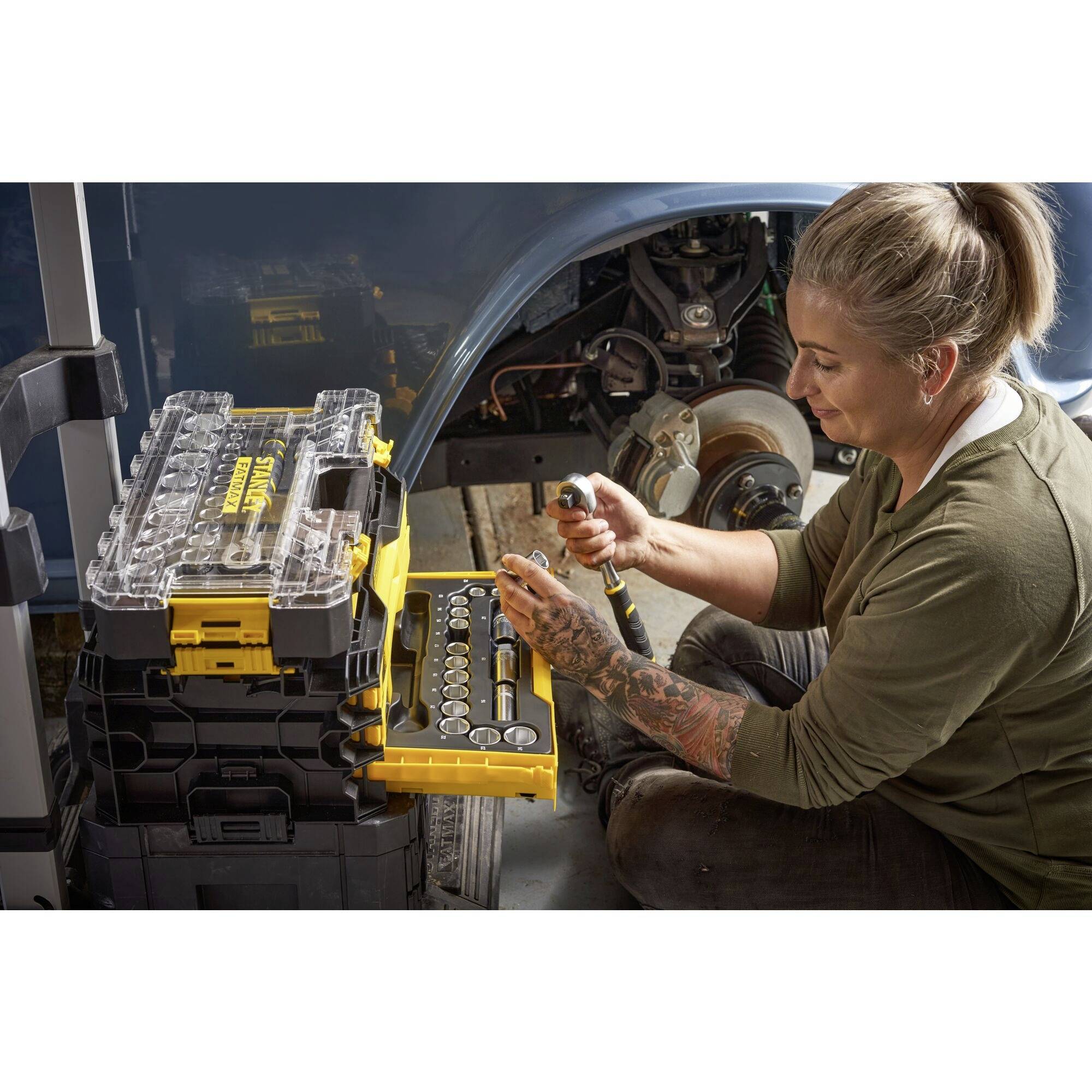 Osoba opravuje automobil, drží v ruce nástroj. V pozadí jsou viditelné montážní skříně.