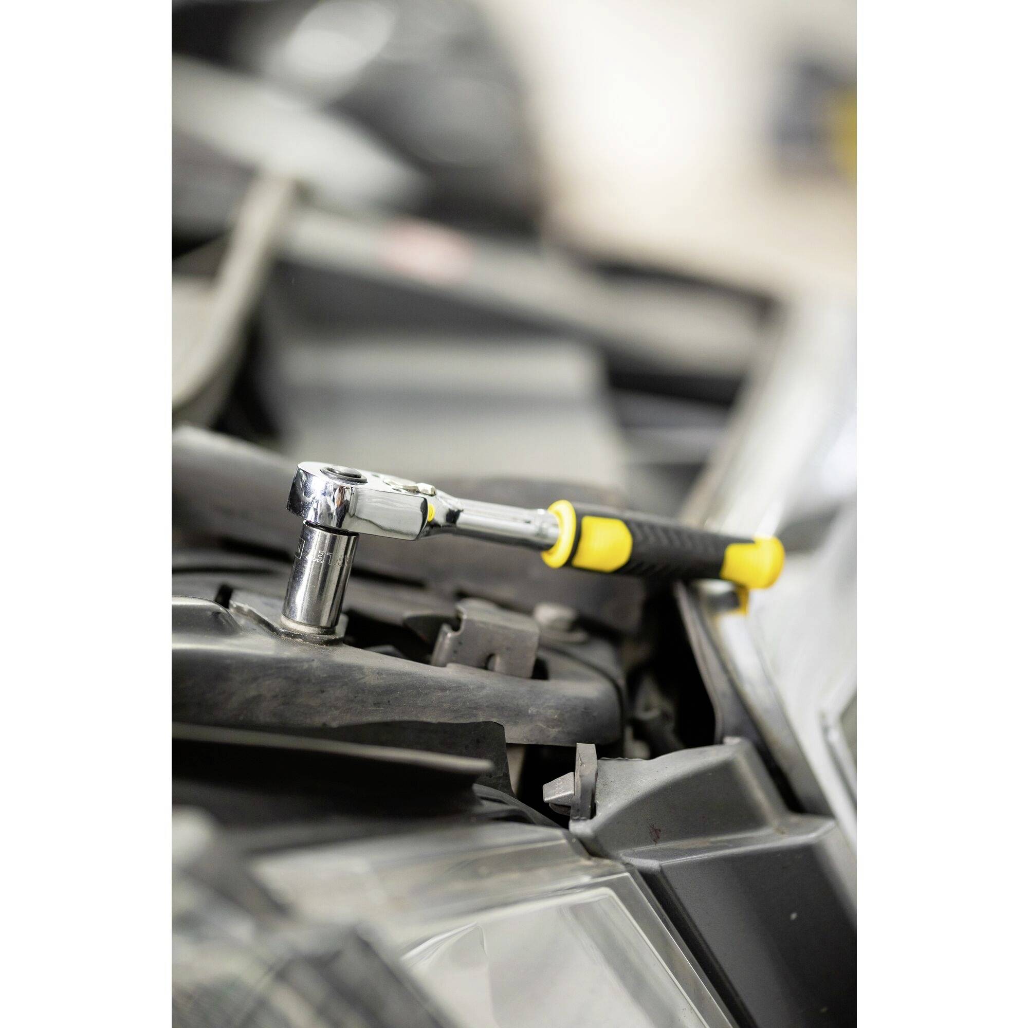 Žlutý klíč leží na autodílu, pravděpodobně v motorovém prostoru vozidla, připravený na opravárenské práce.