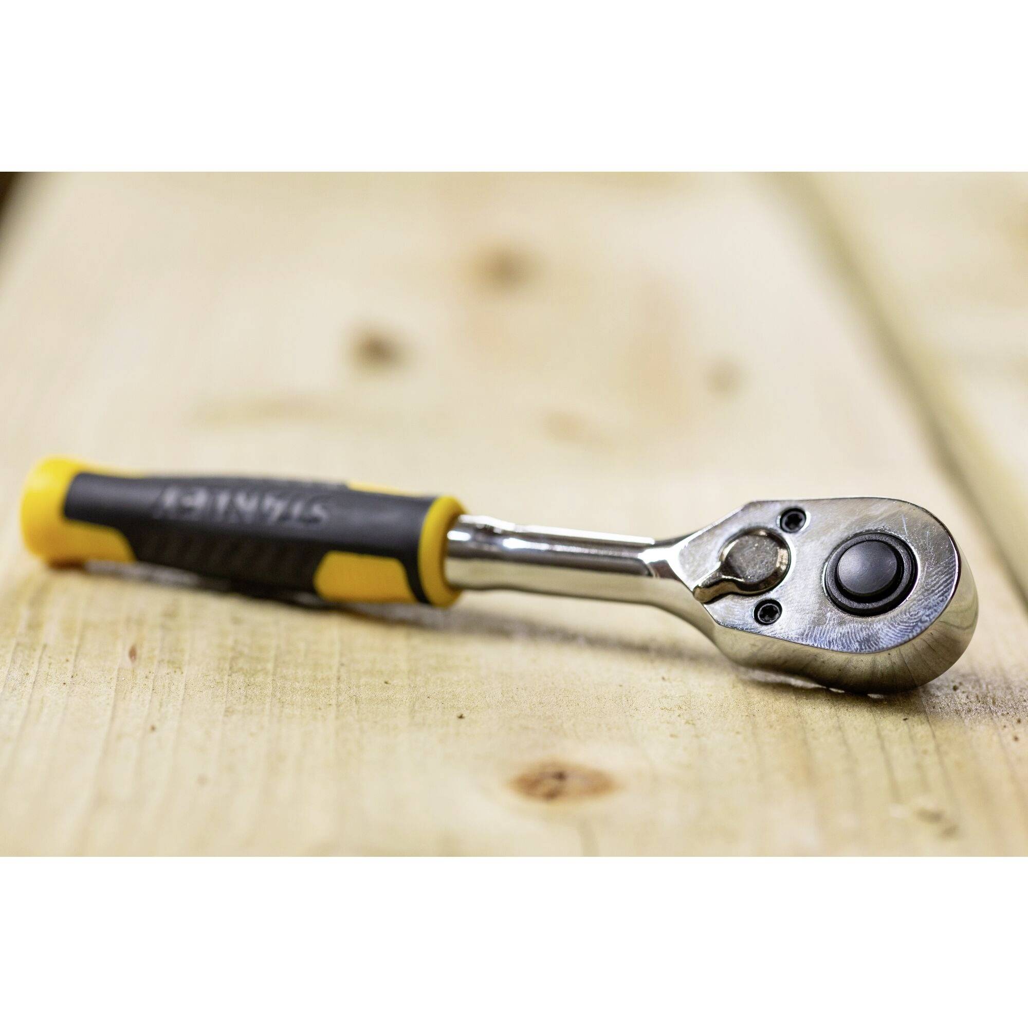 Ráčna leží šikmo na dřevěném stole, s žlutou a černou gumovou rukojetí, stříbrnou hlavou s kulatou pákou, slouží k utahování šroubů.