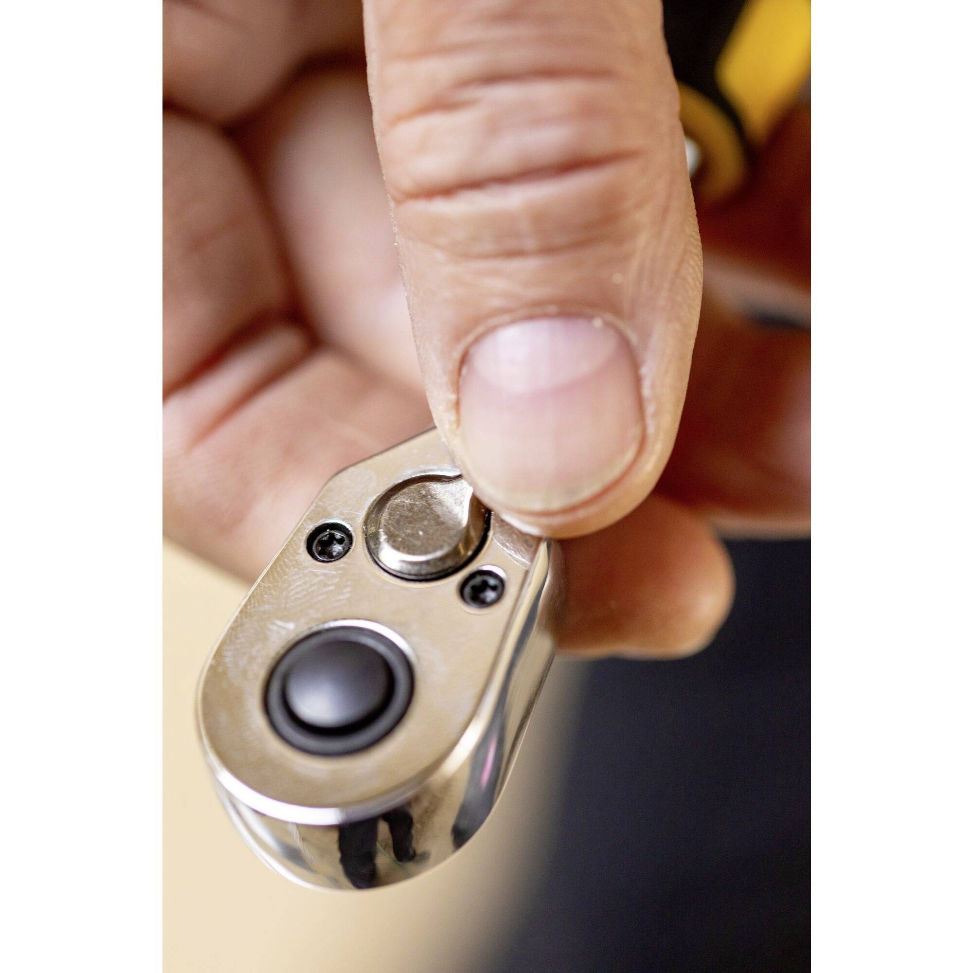 Jedna ruka drží malý kovový spoušťový mechanismus s černým tlačítkem, který pravděpodobně aktivuje nějaký přístroj nebo systém.