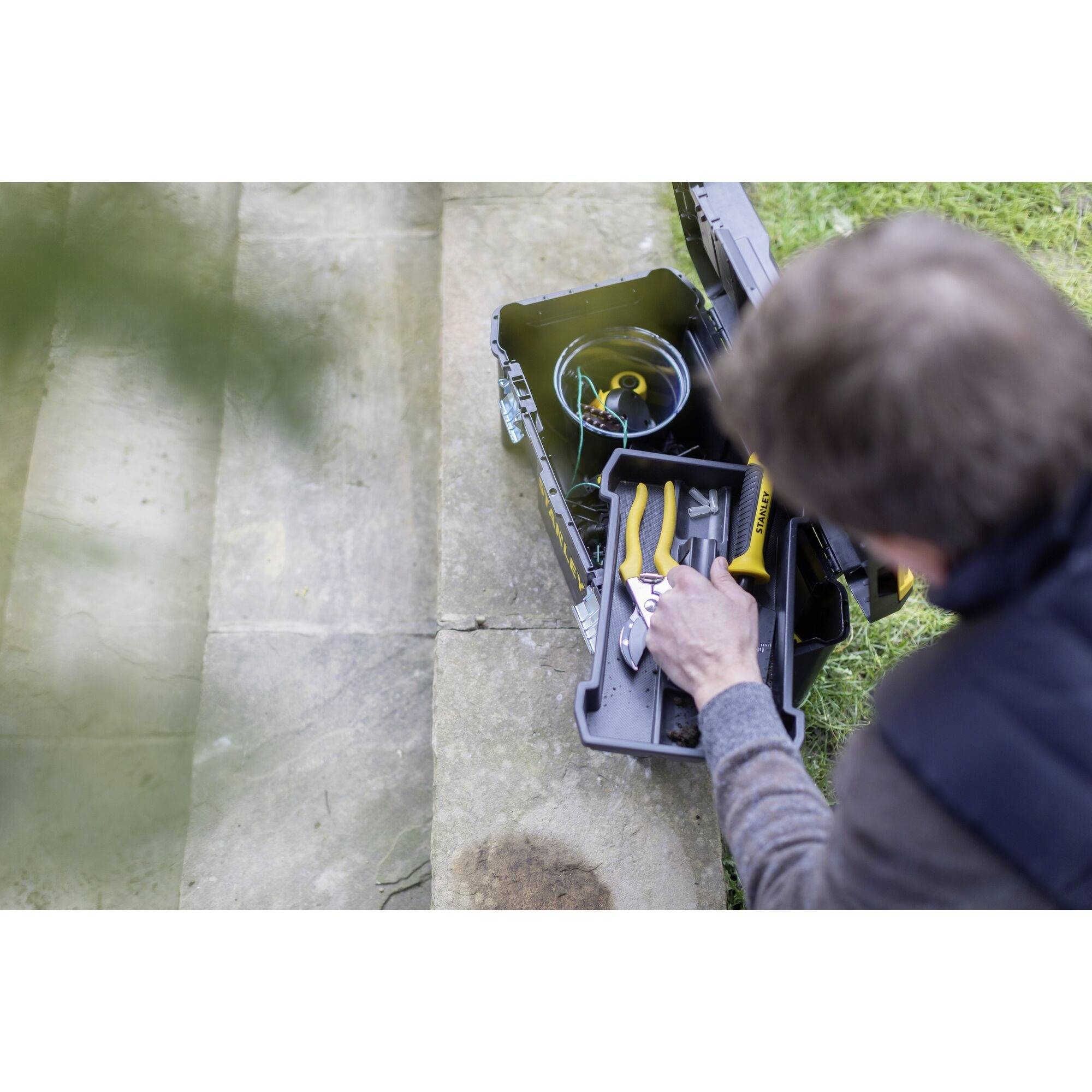 Osoba sahá do otevřené sady nástrojů na terase a vybírá žluté kleště. V pozadí je vidět tráva.