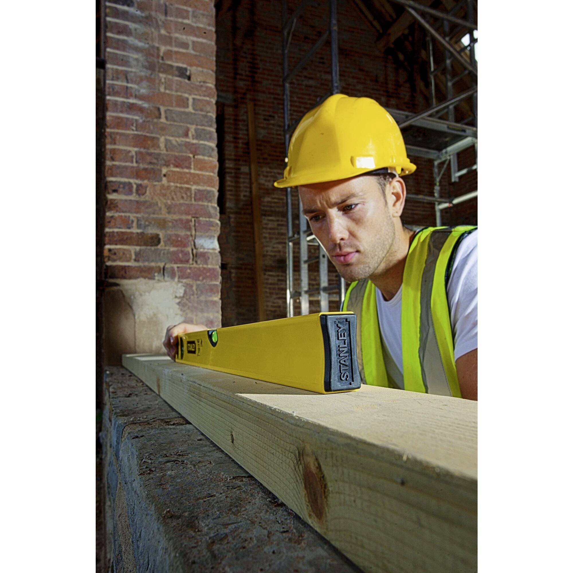 Dělník v přilbě a reflexní vestě kontroluje vodováhou rovnost dřevěného trámu na staveništi.