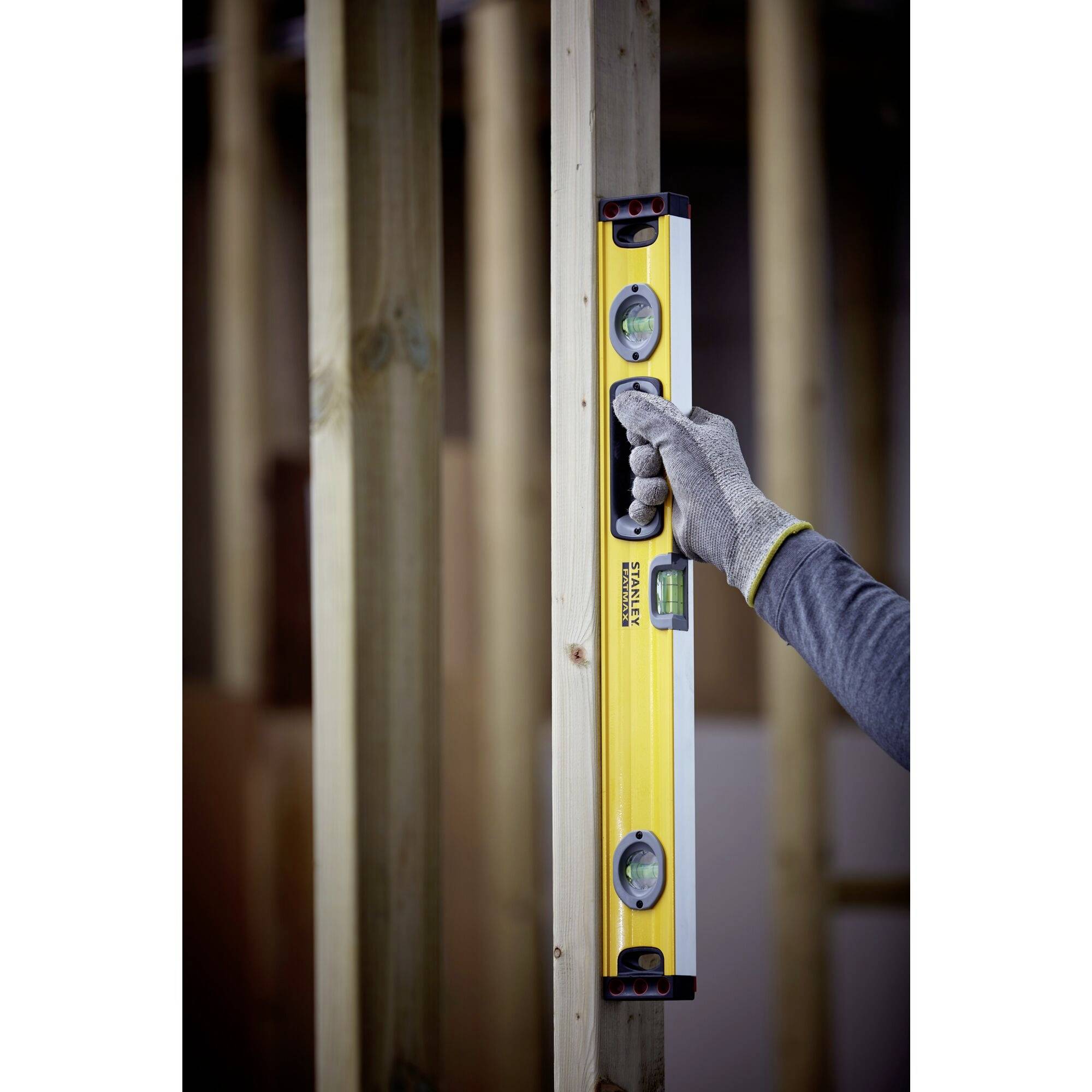 Osoba v rukavici drží žlutou vodováhu svisle u dřevěného kůlu.