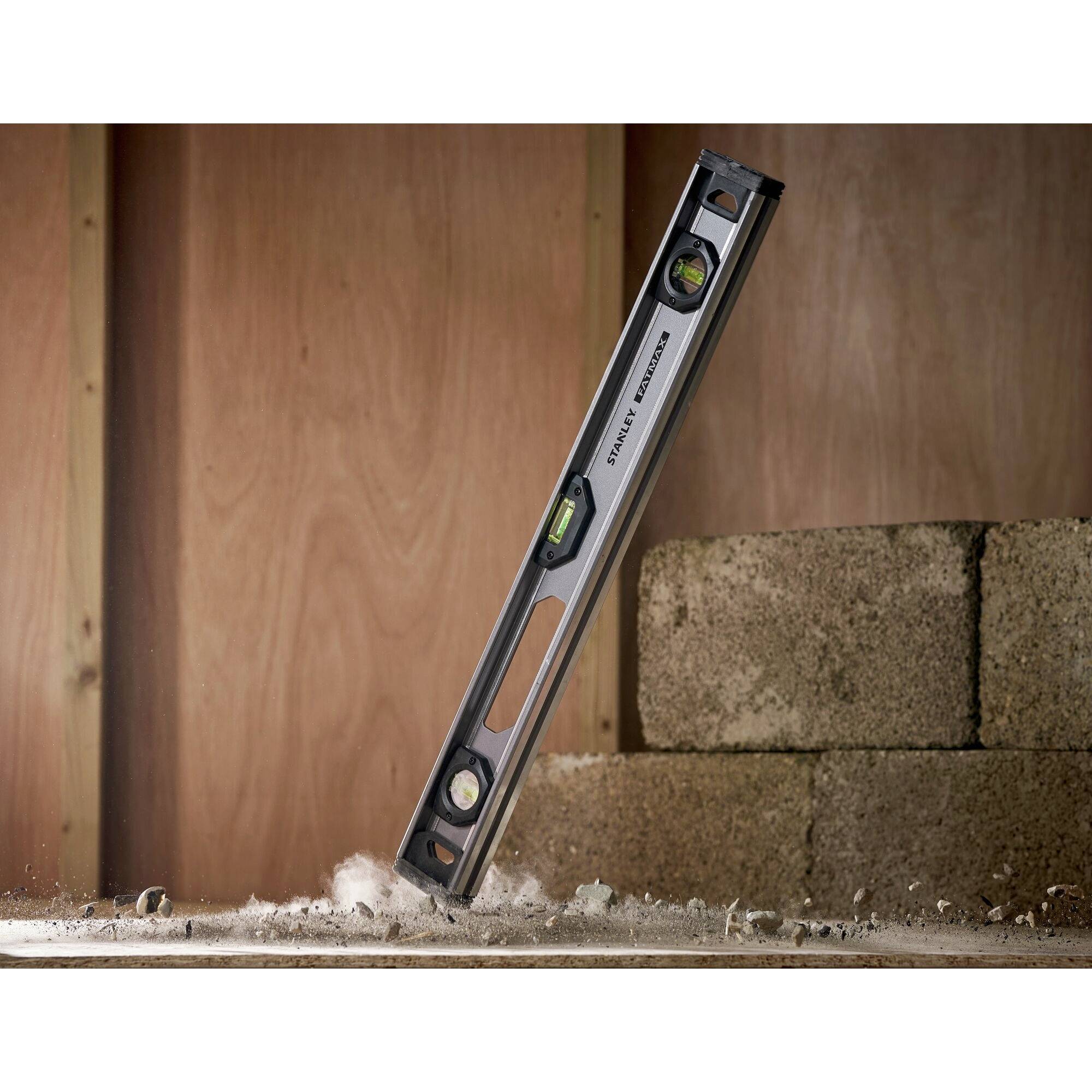 Vodní váha leží diagonálně na dřevěné podlaze, obklopená cihlami a prachem. Osvětlení zvýrazňuje strukturu nástroje.