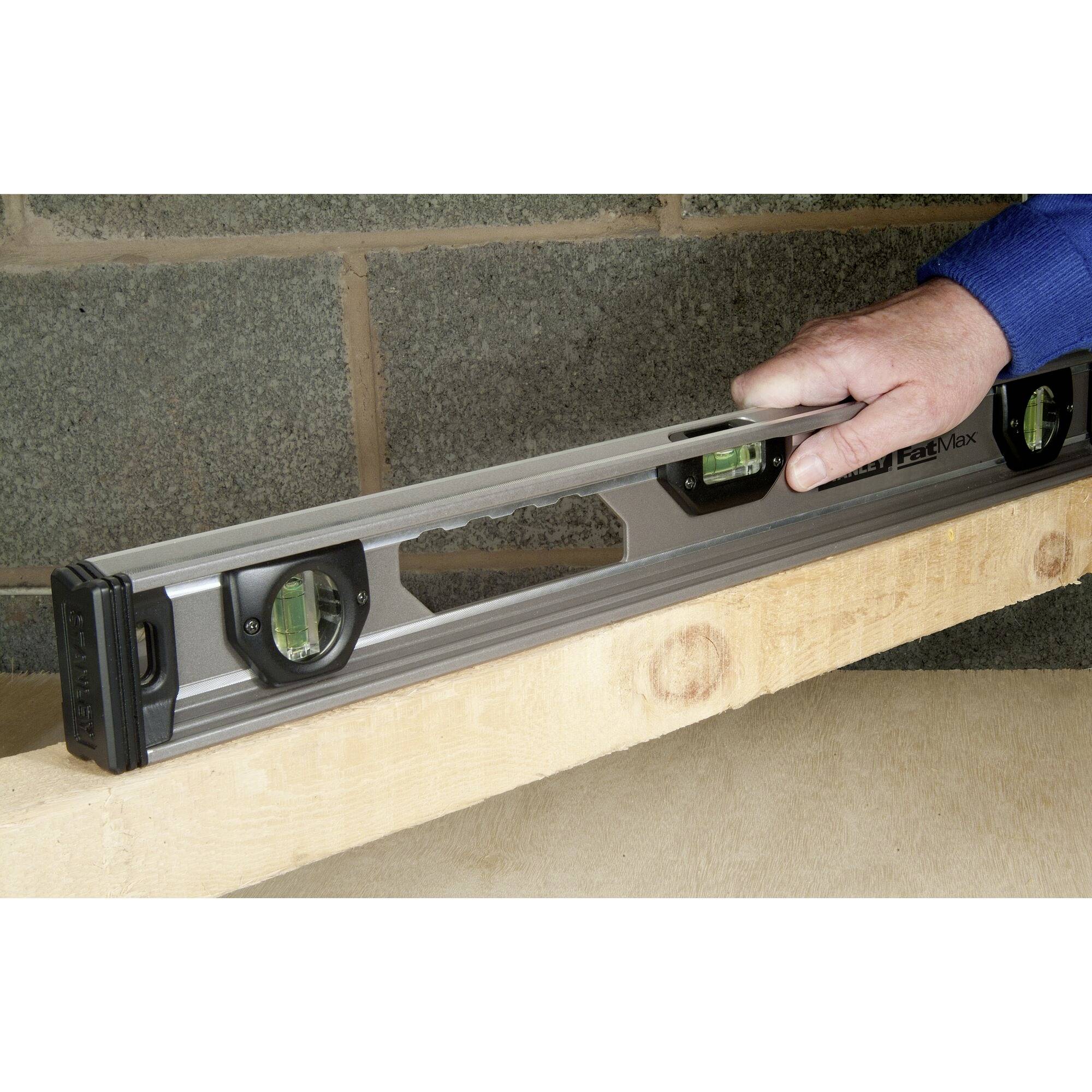 Jedna ruka drží vodováhu na dřevěné desce u betonové zdi. Vodováha ukazuje, zda je deska rovná.