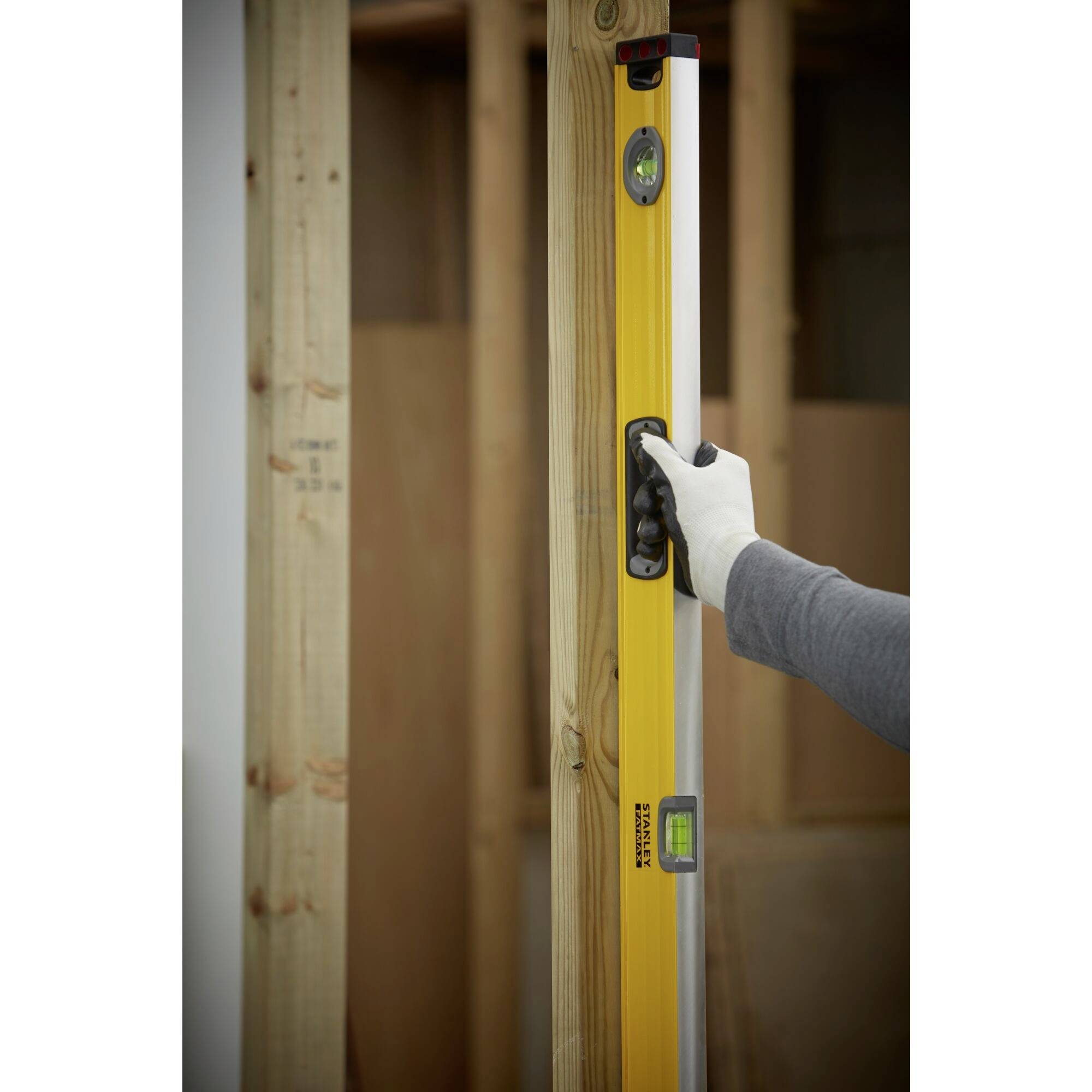 Osoba v pracovních rukavicích přidržuje žlutou vodováhu u dřevěného kůlu, aby zkontrolovala svislou polohu.