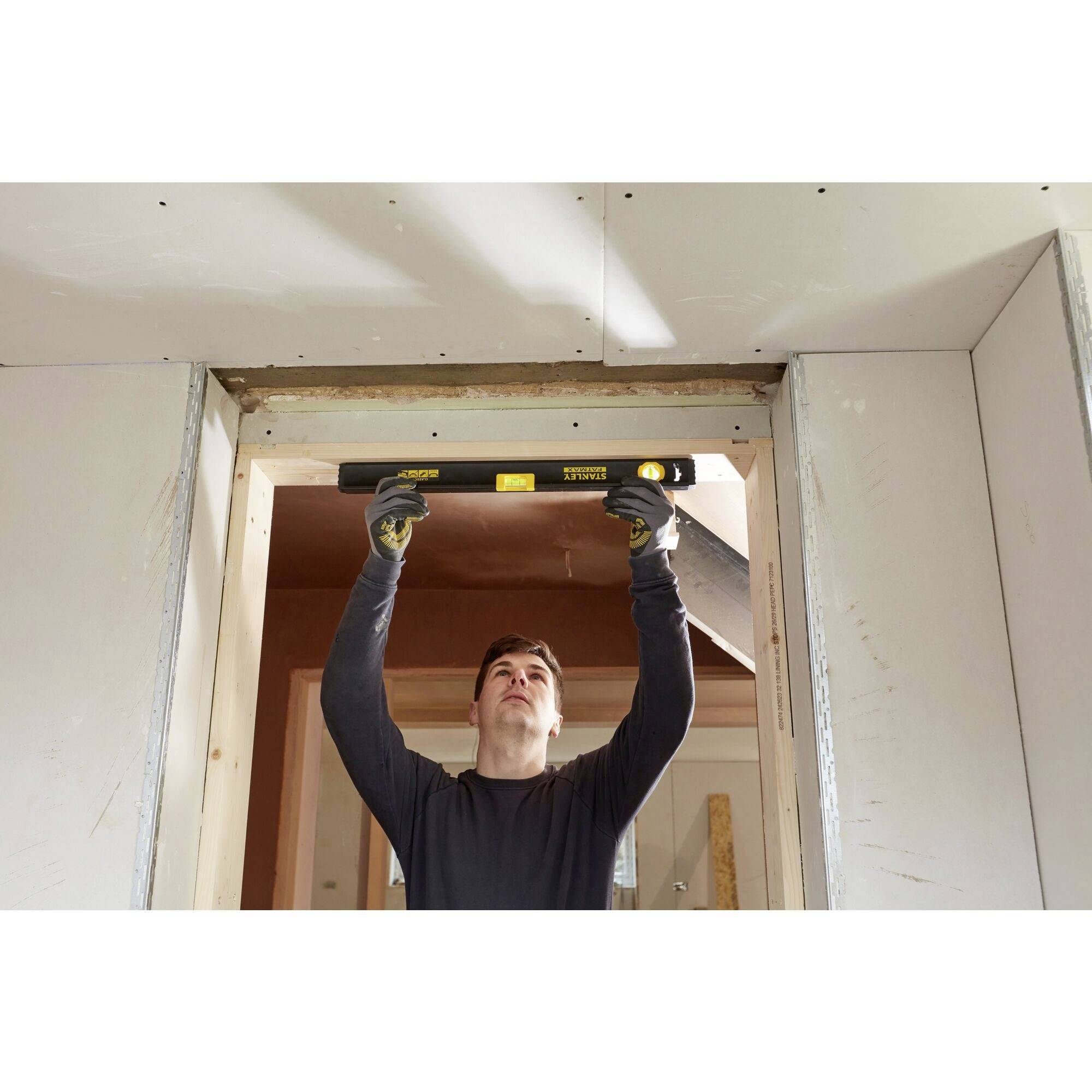 Muž používá vodováhu ke kontrole zárubně dveří. Má nasazené rukavice a pracuje v nedokončené místnosti.