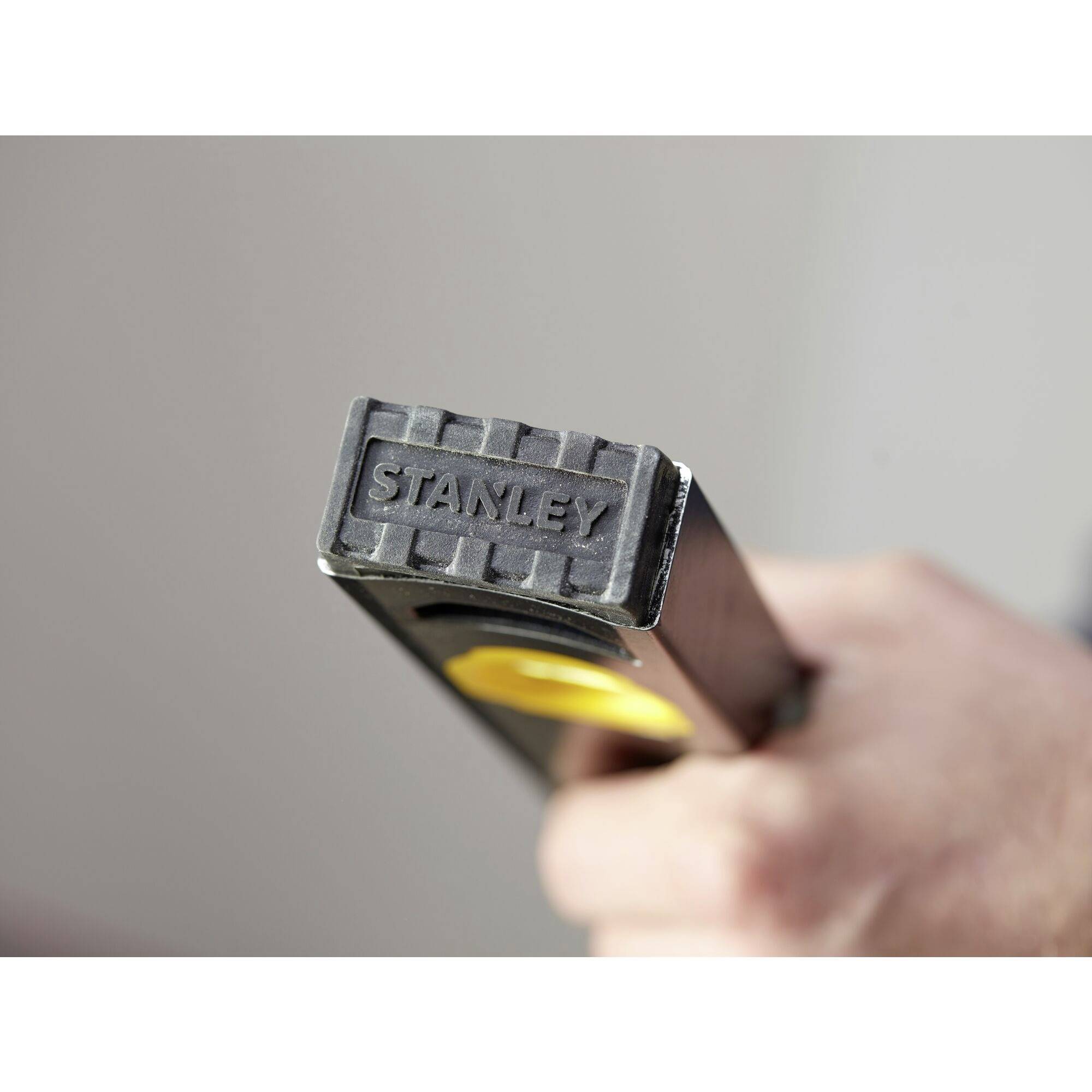 Ruka opatrně drží zblízka stříbrné kladivo Stanley se žlutou rukojetí.