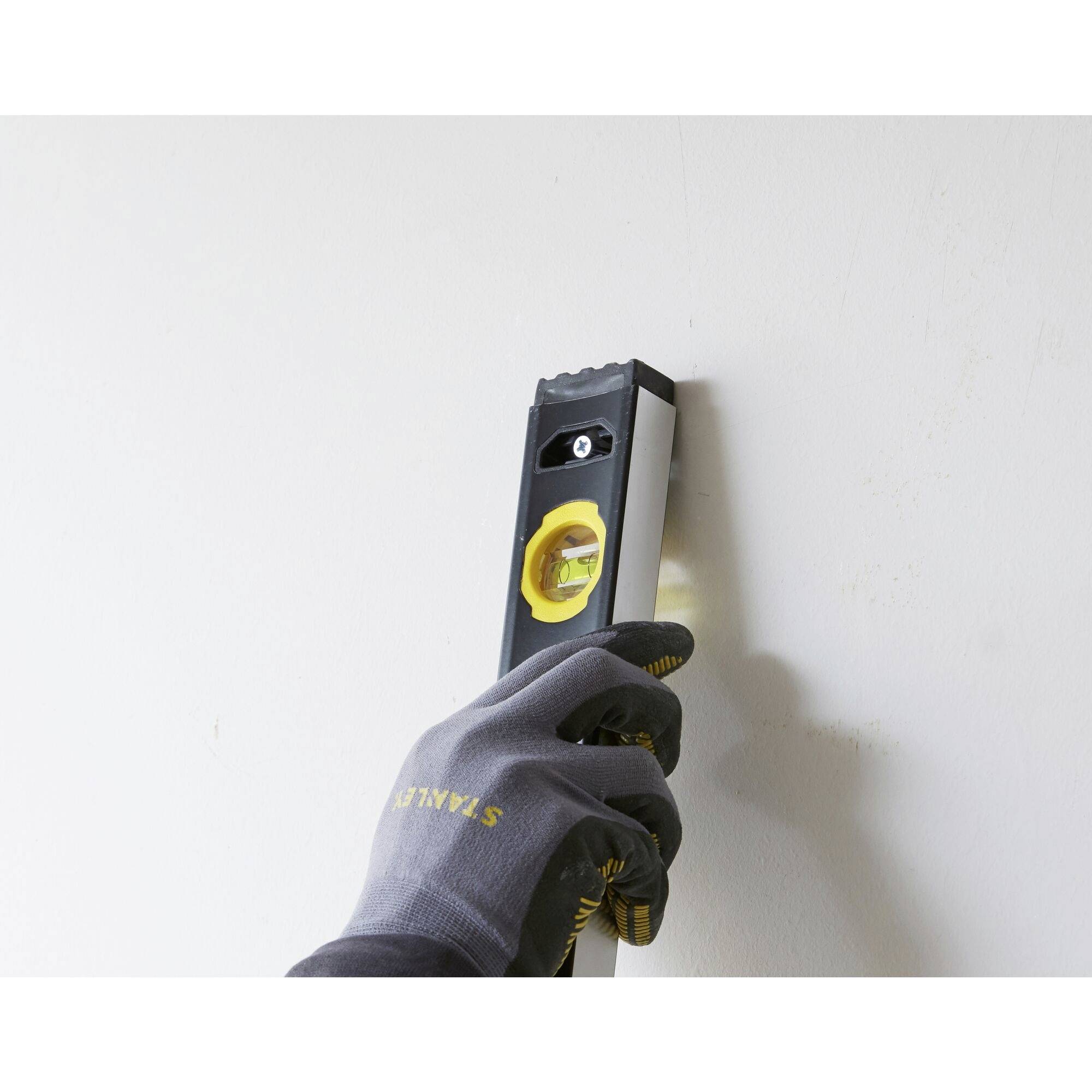 Osoba drží vodováhu u zdi, aby zkontrolovala její rovnost. Osoba má rukavice a zeď je světlá.