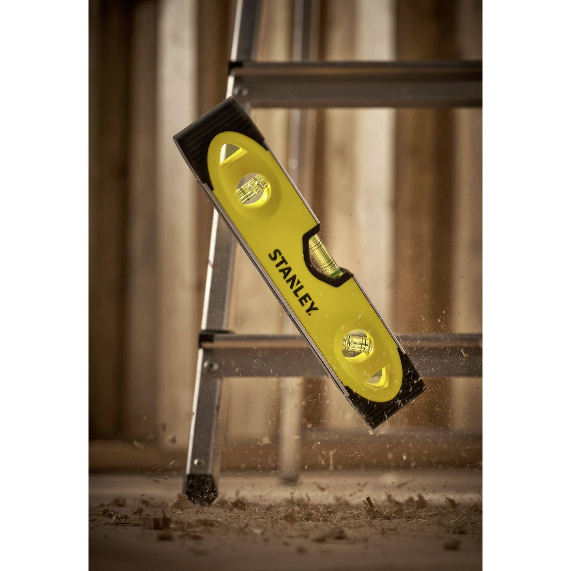 Vodováha padající z žebříku na zem, obklopená dřevěnými pilinami, symbolizuje nebezpečí úrazu při stavebních pracích.