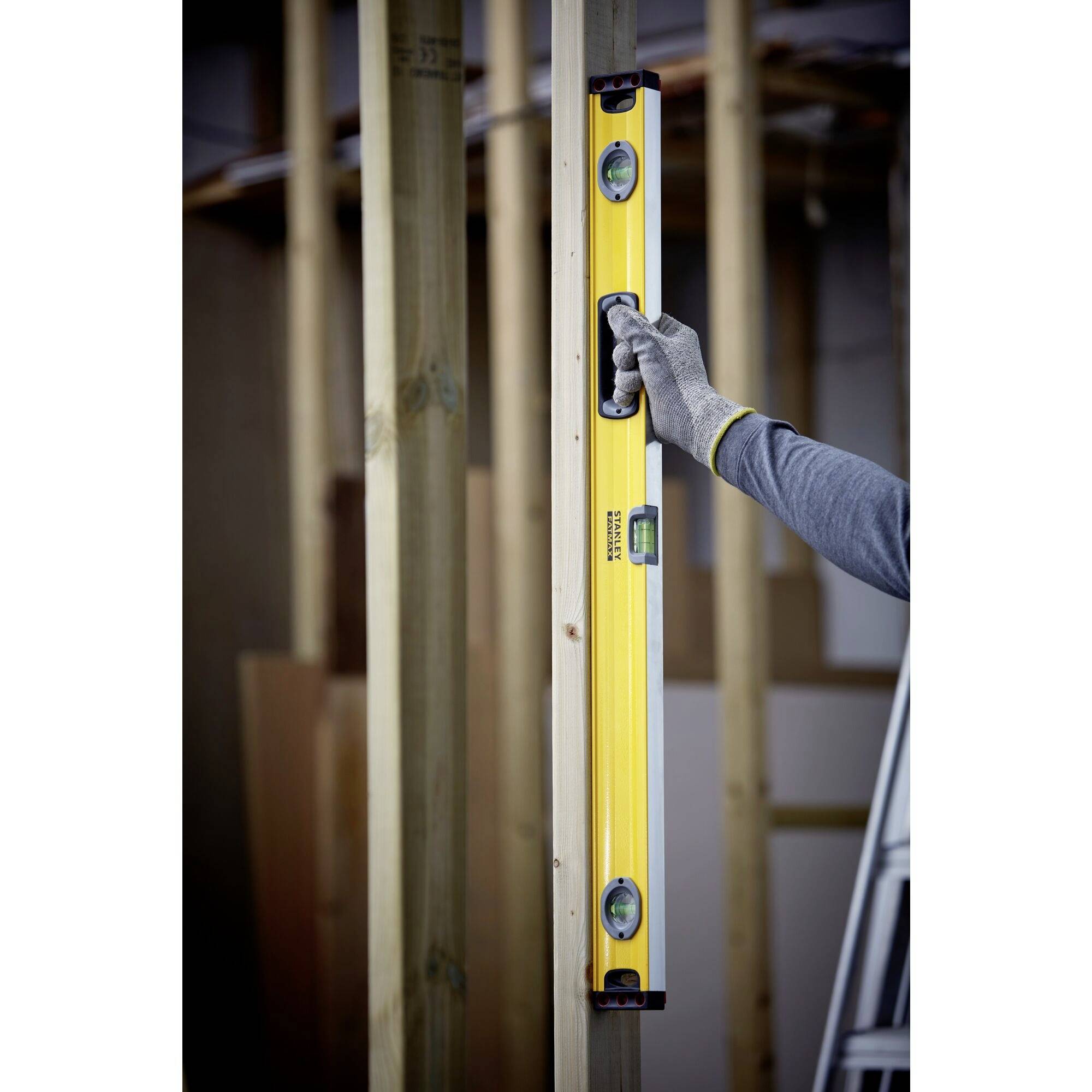 Osoba drží žlutou vodováhu svisle u dřevěného sloupku, aby zkontrolovala svislost. V pozadí jsou vidět další dřevěné trámy.