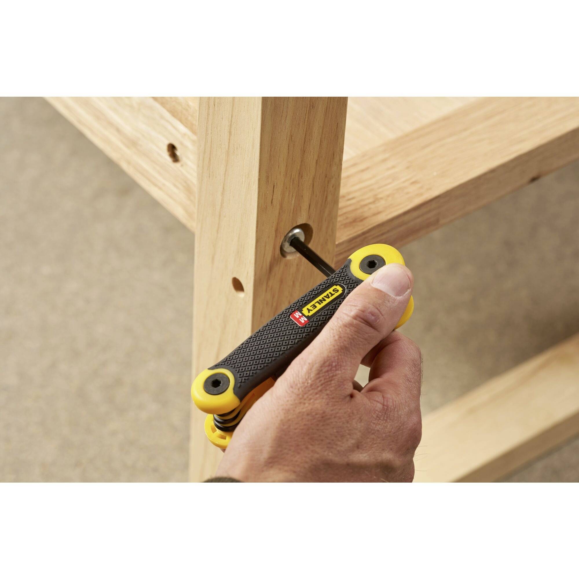 Jedna ruka utahuje šroub žlutým vícenásobným šestihranným klíčem na dřevěné polici.