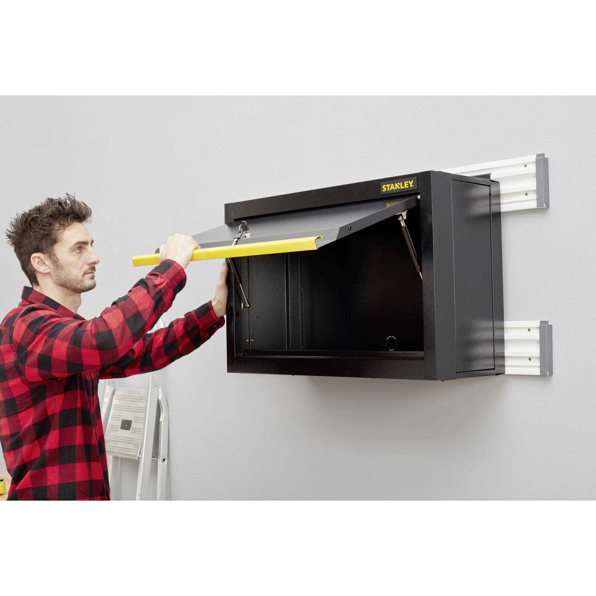 Muž otevírá černou nástrojovou skříň připevněnou na stěně se žlutou rukojetí v dílně. Za ním stojí žebřík.