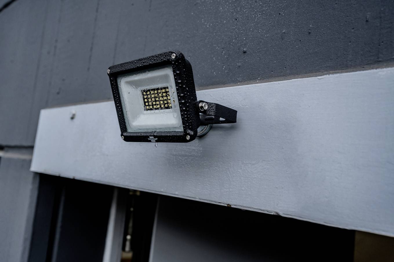 LED reflektor na šedé stěně, pravděpodobně určený pro venkovní osvětlení, s kapkami vody na povrchu.