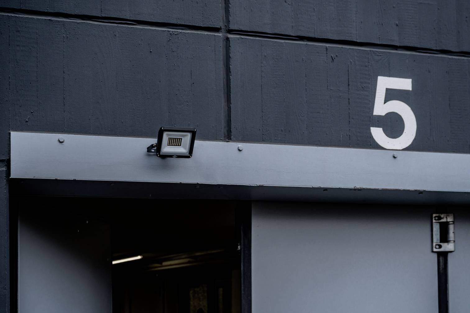 Šedá stěna s velkou bílou číslicí 5. Pod ní jsou otevřené dveře a malý reflektor.