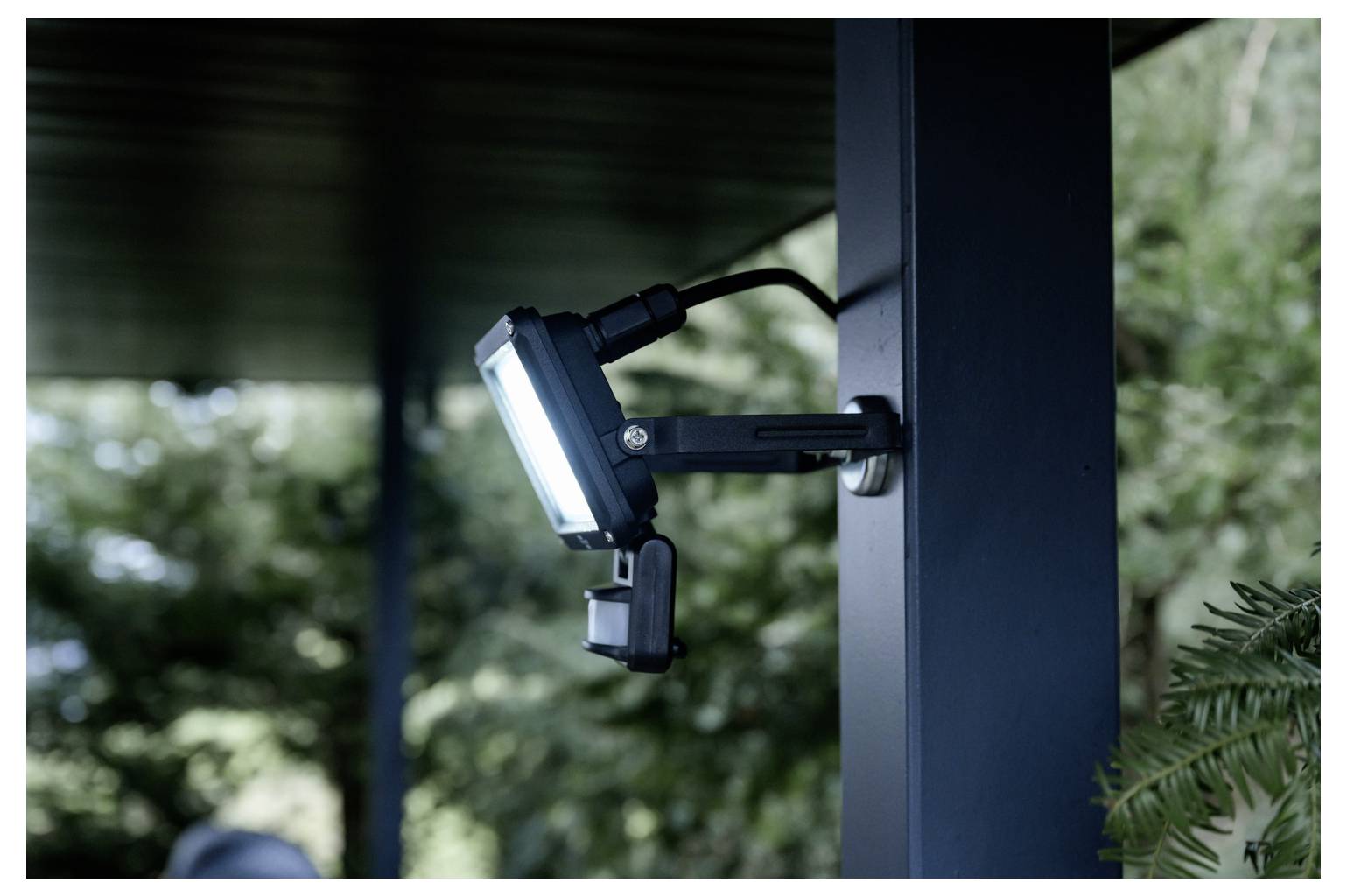 Připevněné LED bezpečnostní světlo na tmavém sloupu pod střechou, zaměřené směrem ven do exteriéru s rozmazanými stromy v pozadí.
