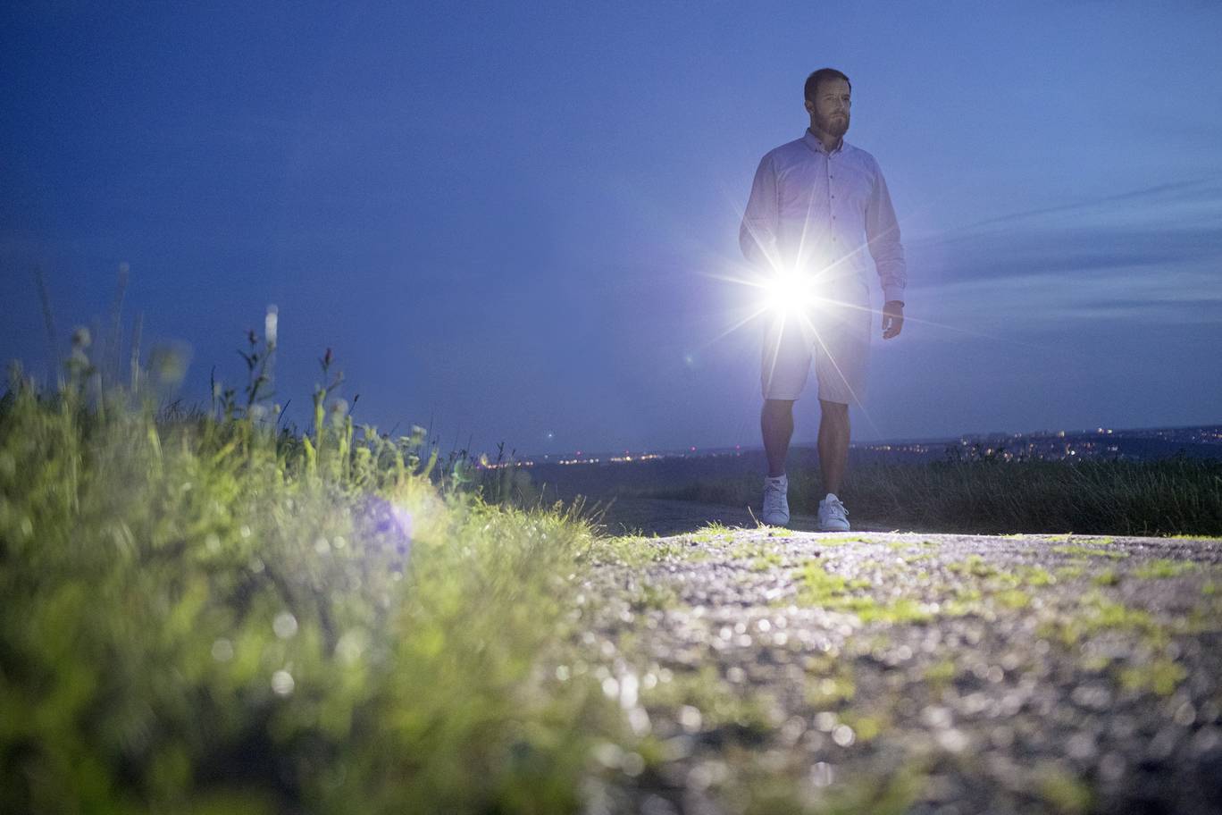 Osoba v noci s baterkou na kamenité cestě, obklopená trávou, v pozadí rozmazané městské světlo.