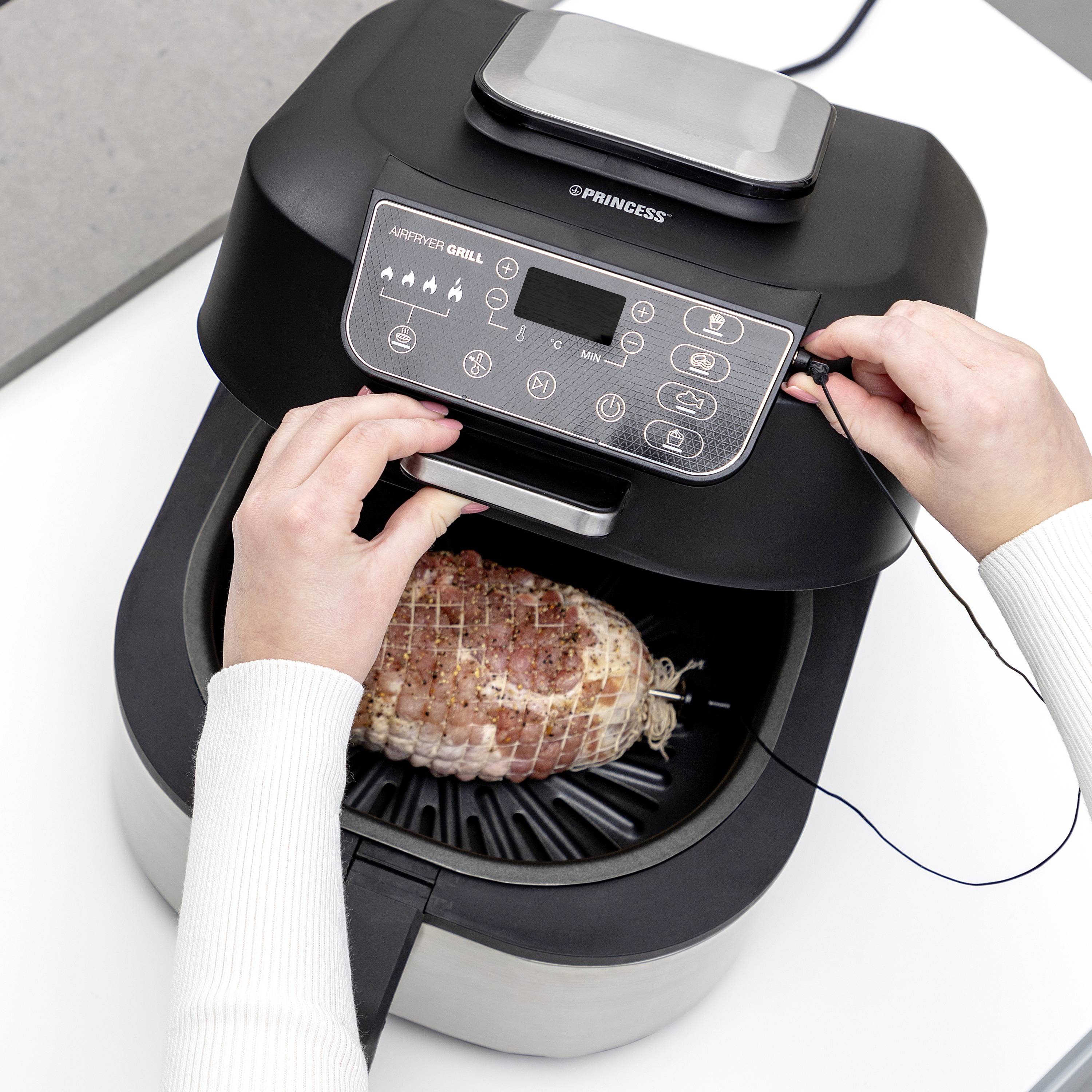 Osoba vkládá kus masa do horkovzdušné fritézy. Ovládací panel horkovzdušné fritézy je viditelný.
