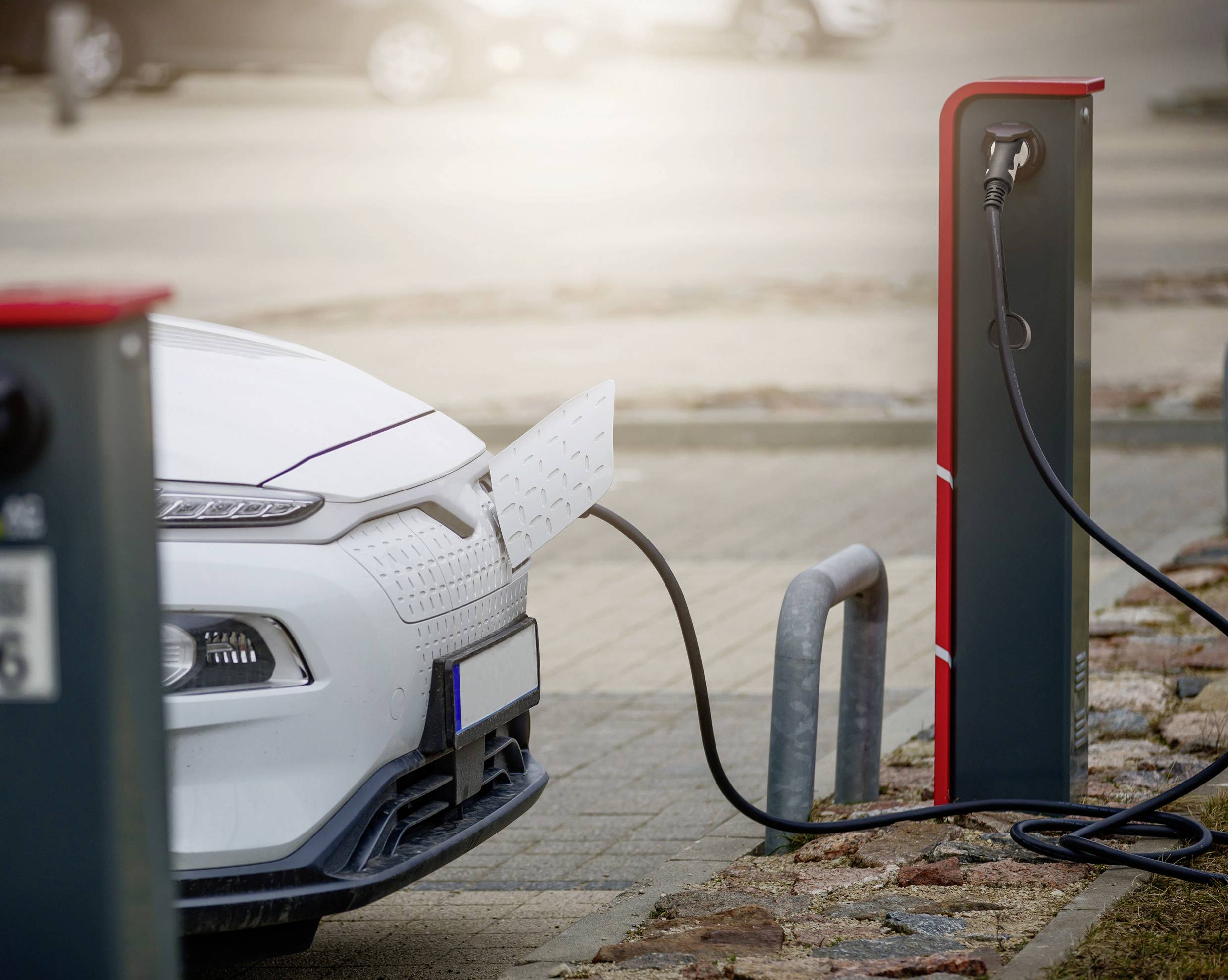 Bílé auto se nabíjí na dobíjecí stanici pro elektromobily. Kabel propojuje stanici s nabíjecím portem vozidla.
