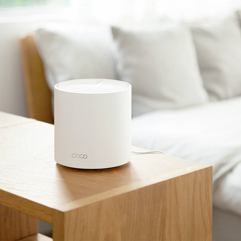 Malý bílý Wi-Fi router stojí na dřevěném stole v světlé místnosti s pohovkou v pozadí.
