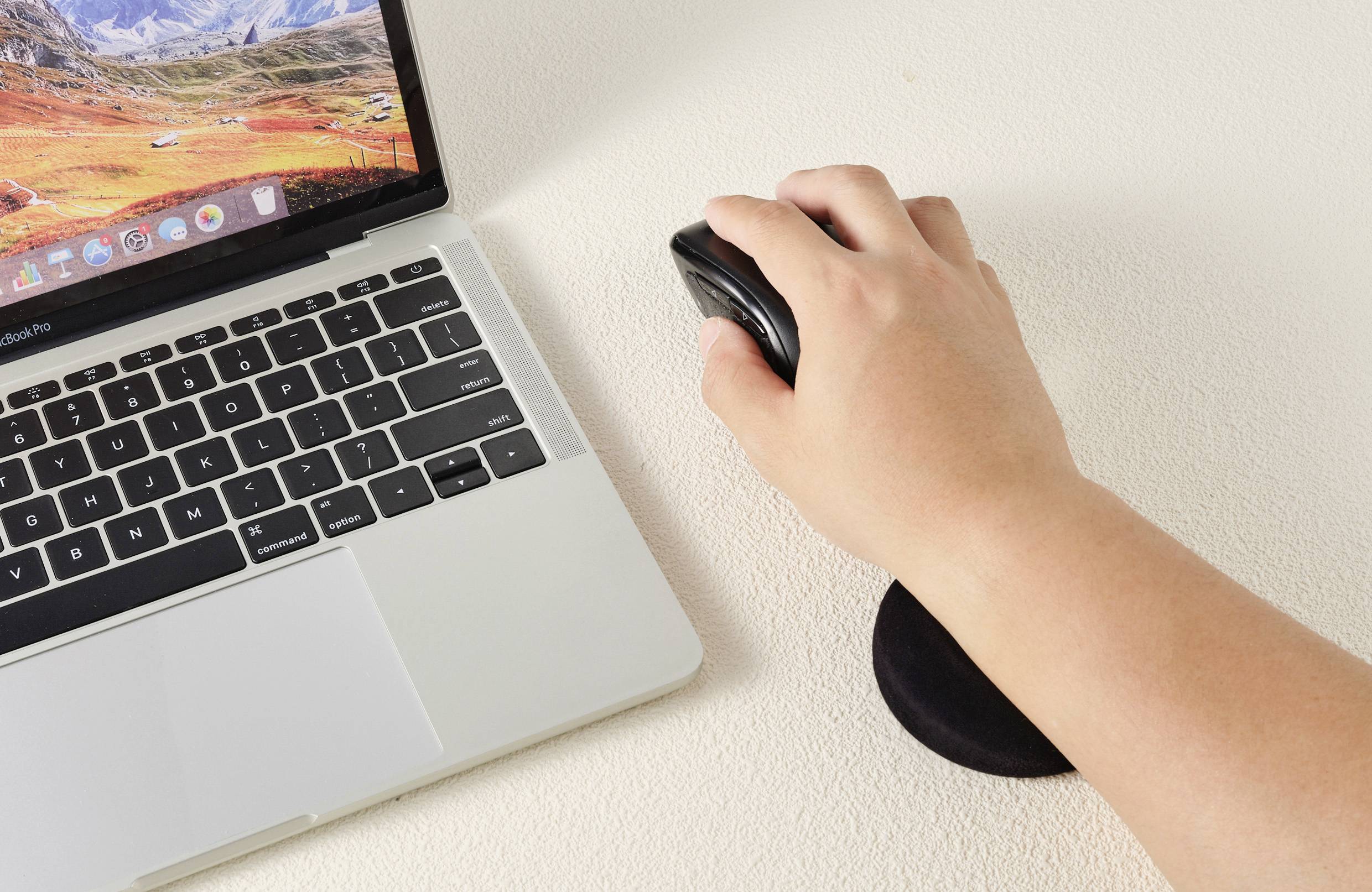 Ruka používá myš vedle otevřeného laptopu na pracovním stole. Laptop zobrazuje krajinu.