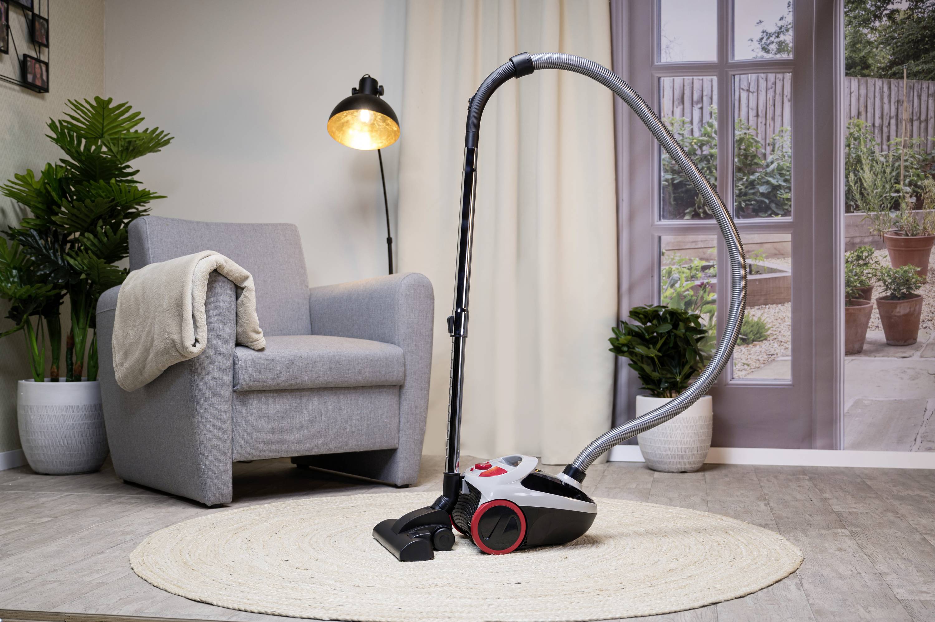 Vysavač stojí v útulném obývacím pokoji vedle křesla na koberci. V pozadí jsou rostliny a lampa.