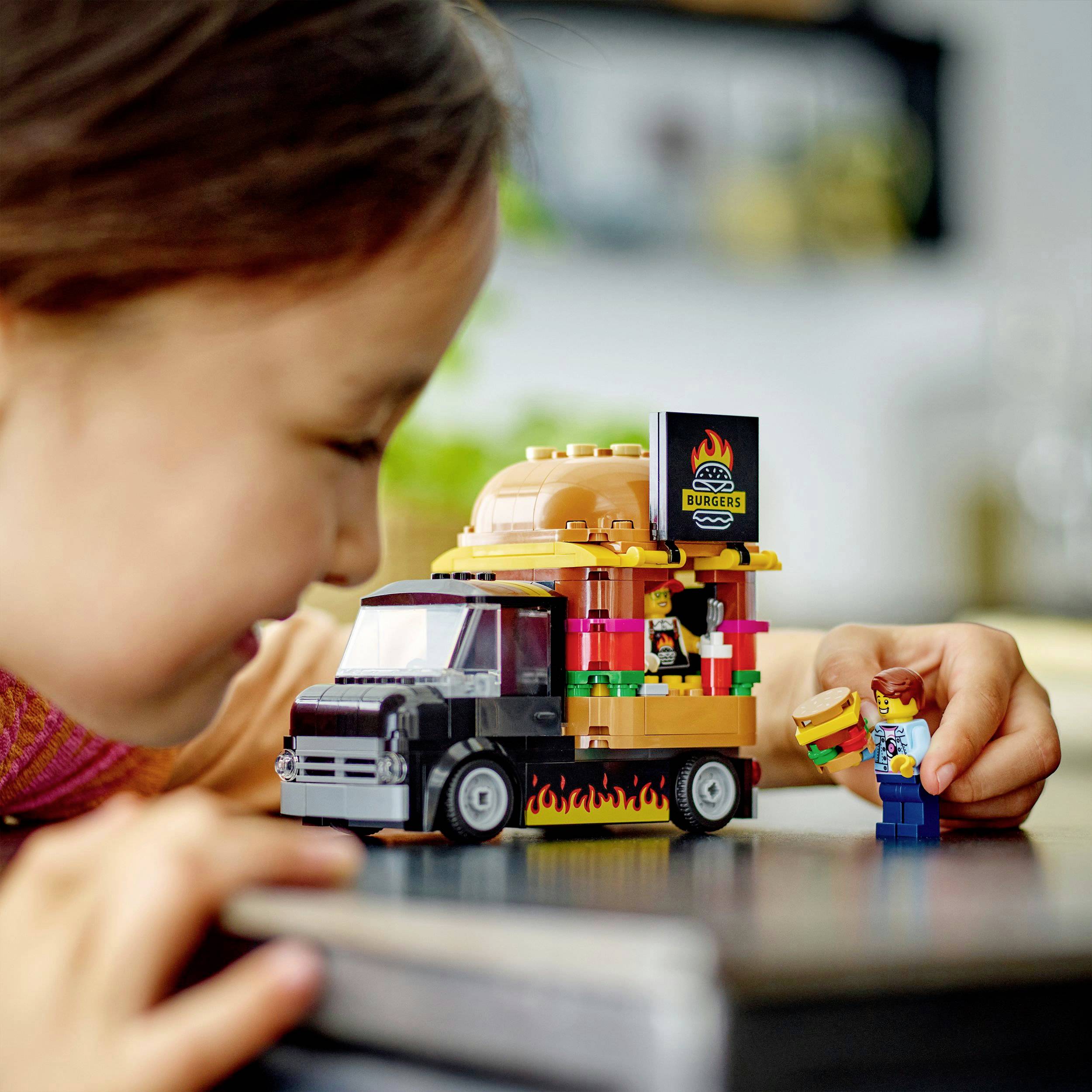 Dítě šťastně staví s LEGO stavebnicí náklaďák s hamburgery, včetně figurky muže s hamburgerem.